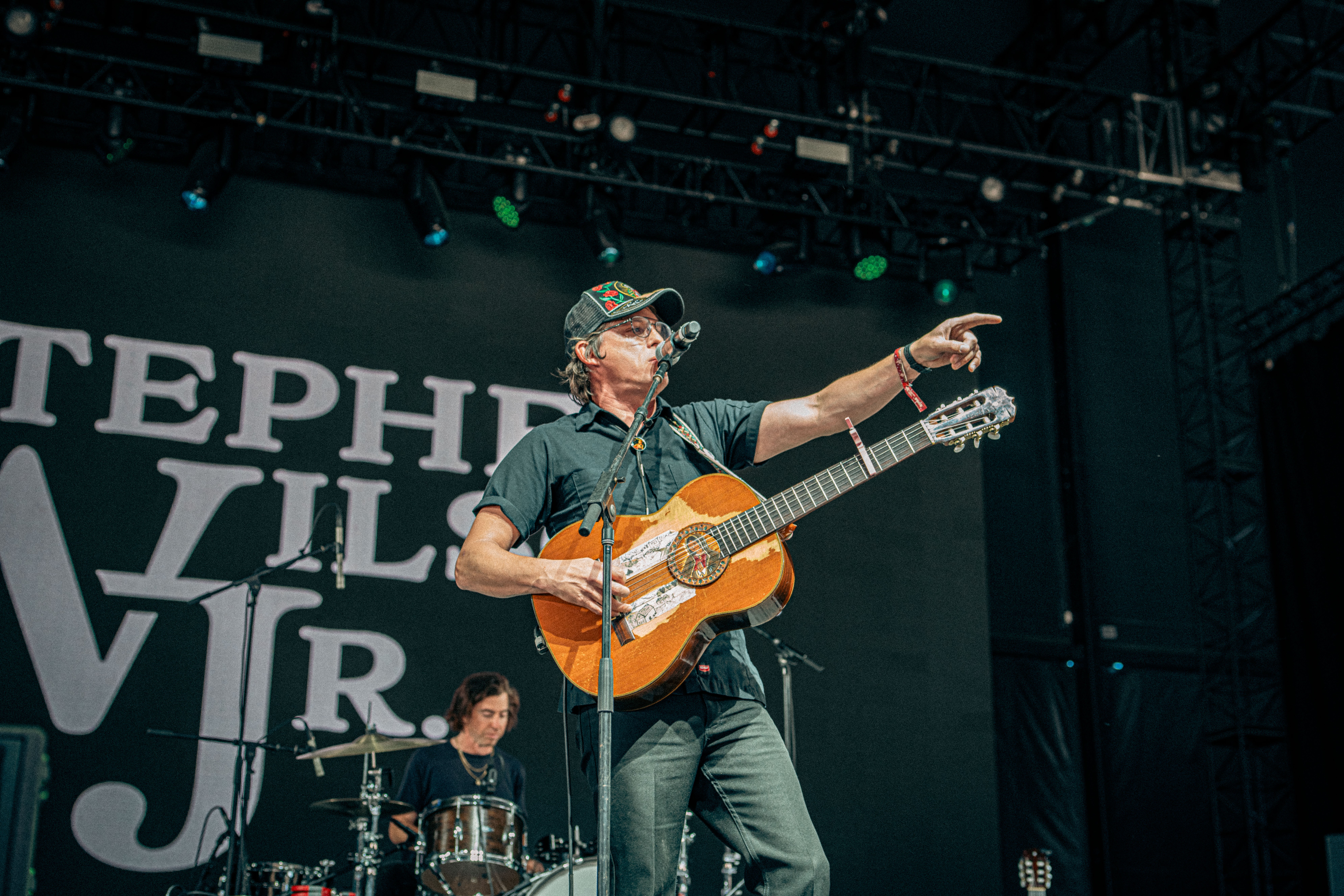 Musician with a guitar points towards the audience on stage, with a drummer in the background and a large screen.