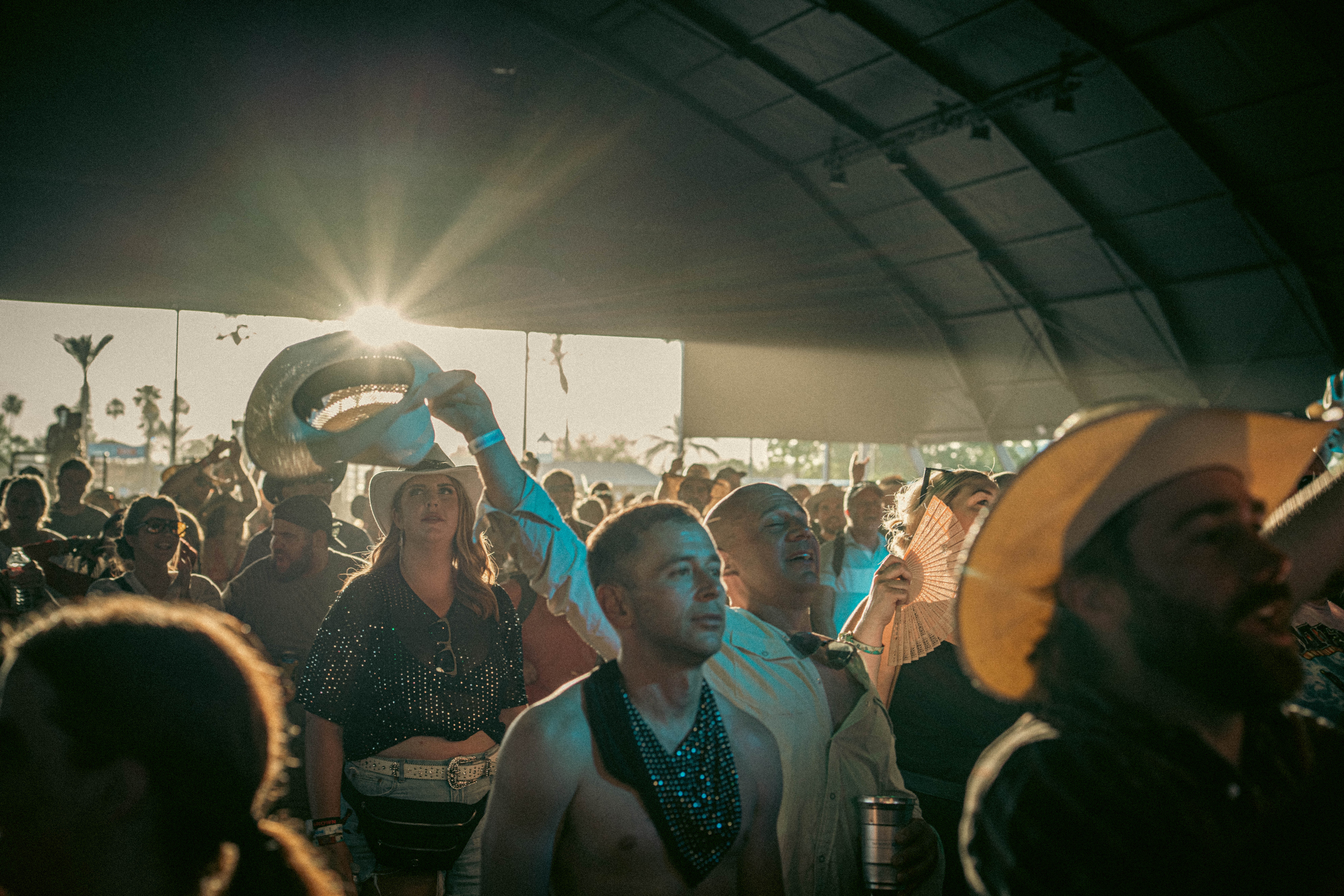 Crowd of people inside a tent with sunlight shining through, some holding umbrellas and taking photos.