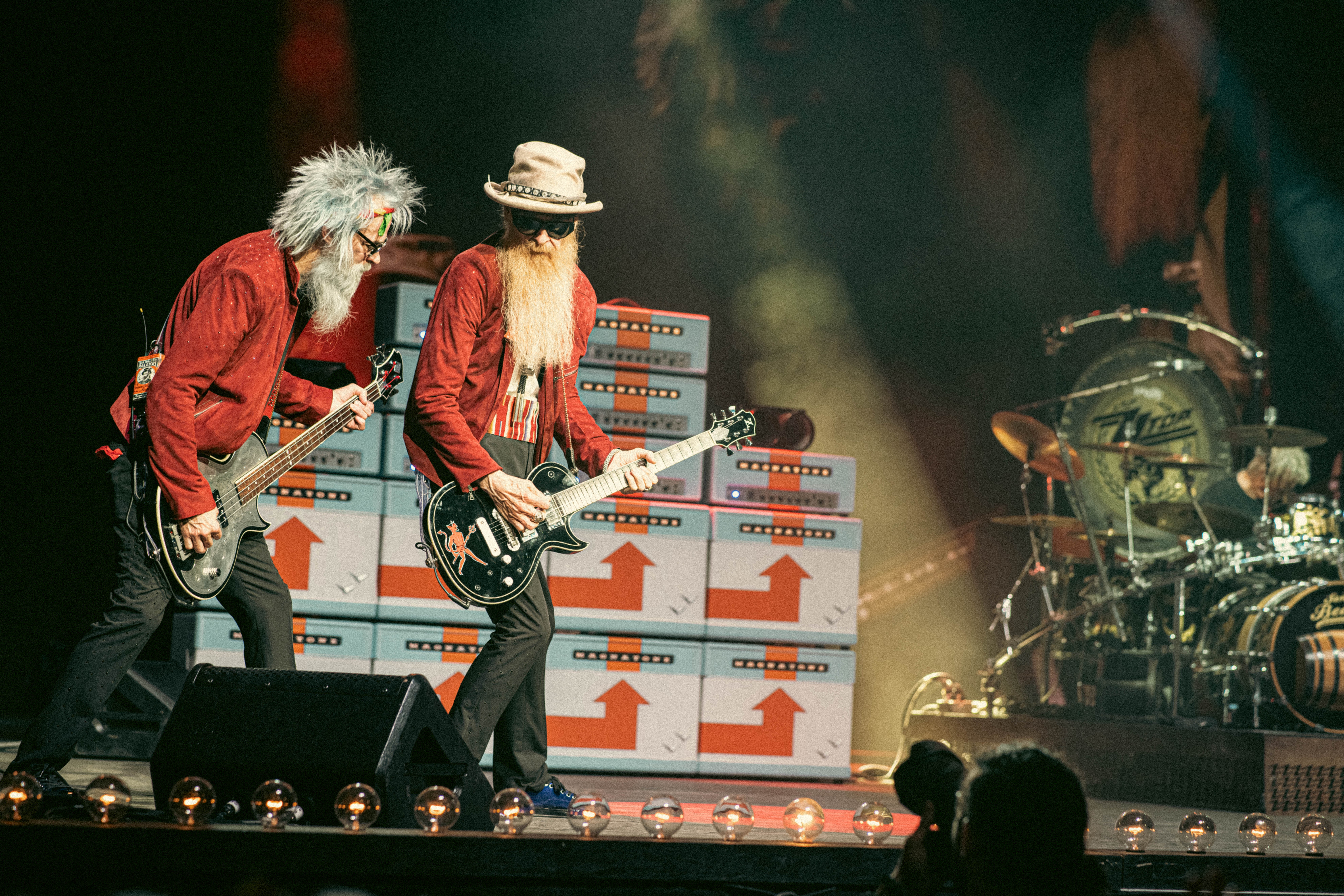 Three musicians perform on stage with guitars and drums, colorful stacked amplifiers in the background, stage lights shining down.