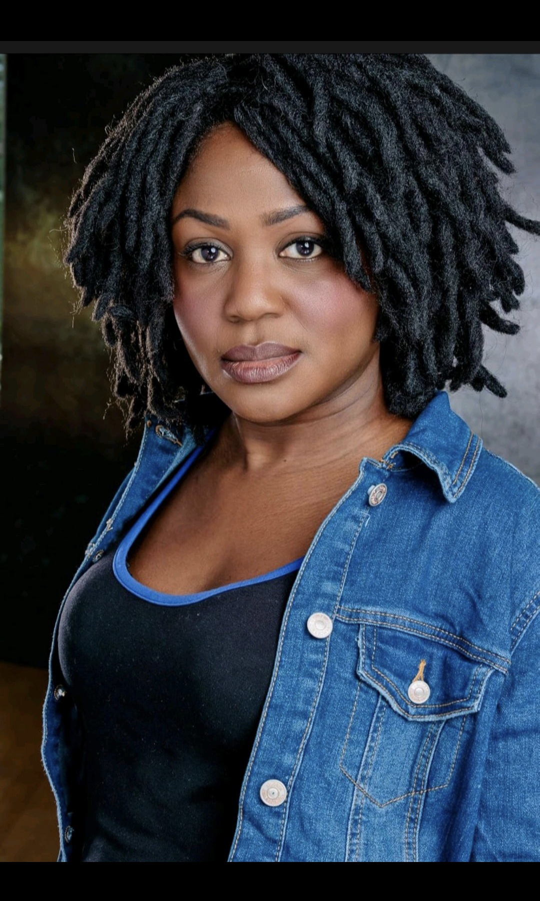 Woman with natural curly hair wearing a denim jacket and black top, looking at the camera.