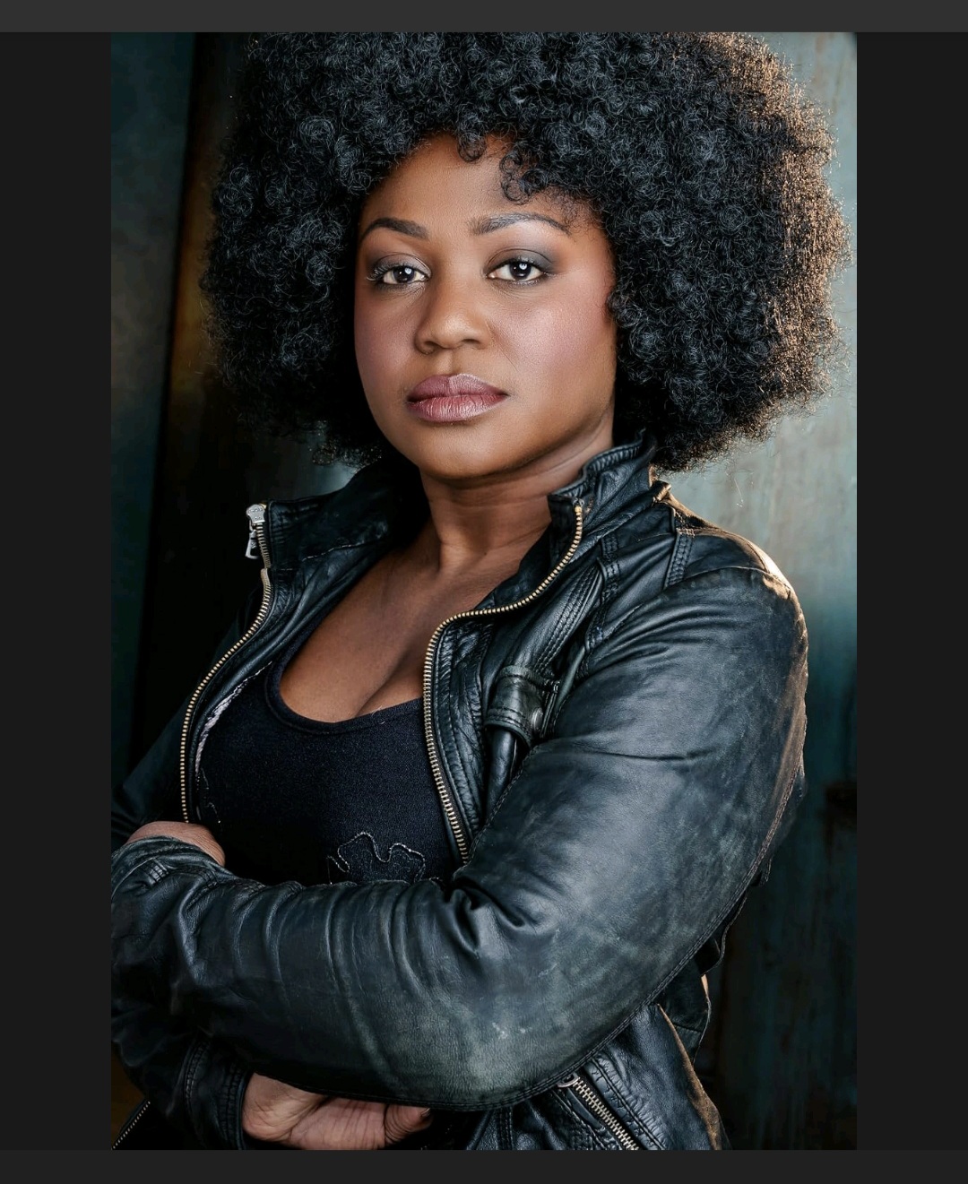 Woman with curly hair wearing a black leather jacket, arms crossed, looking at the camera.
