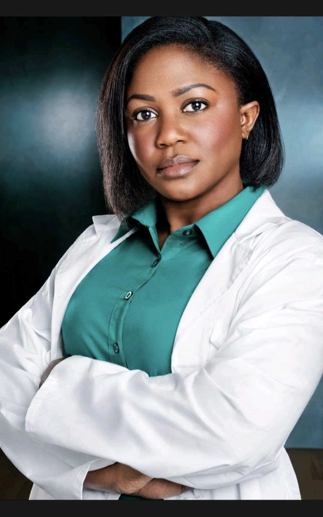 A woman with shoulder-length black hair wearing a teal shirt and white lab coat, arms crossed, looking at the camera.