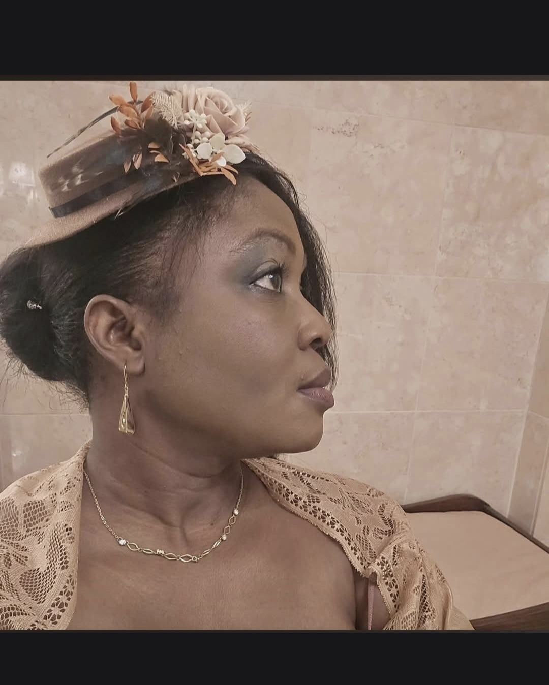 Side profile of a woman wearing a hat with flowers, earrings, and a lace top, against a plain background.