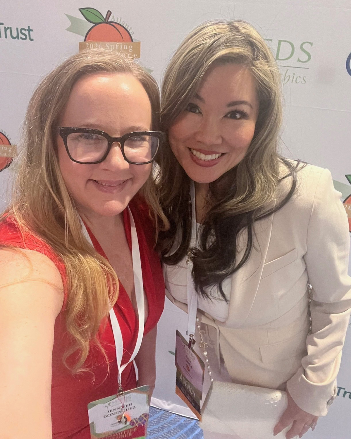 Two women smiling, one with glasses and red top, the other with long hair and white blazer, standing close together.