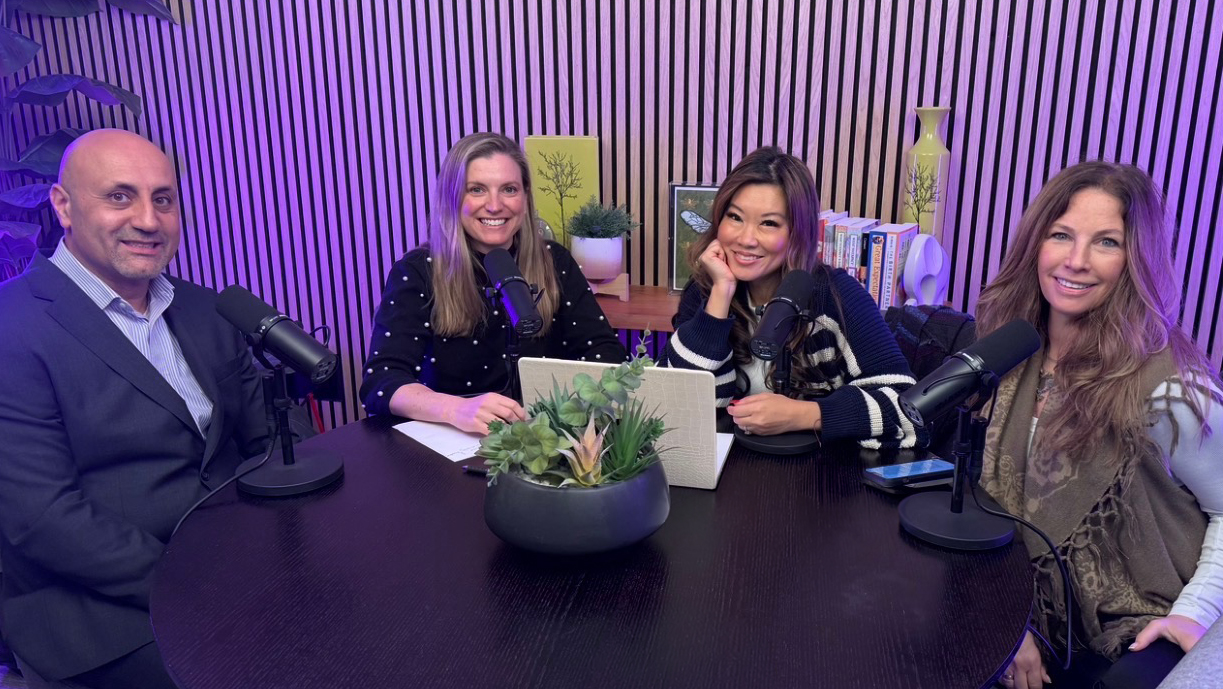 Four people sit at a table with microphones, smiling, in a room with purple lighting and striped wall background.