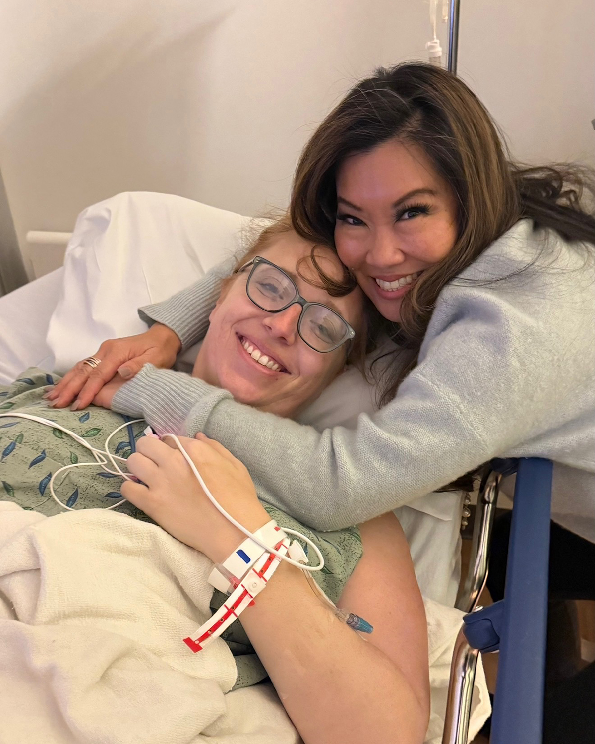 Two women smiling, one lying in a hospital bed with medical wires, the other hugging her, both happy.