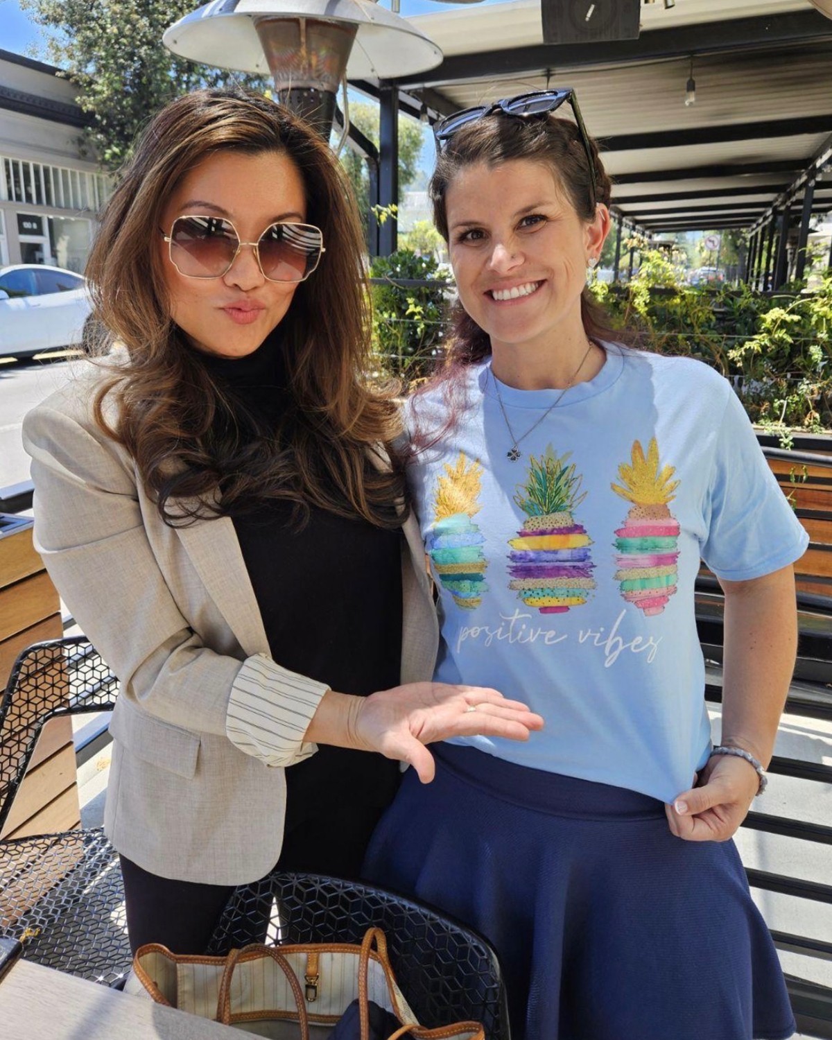 Two women smiling outdoors, one wearing sunglasses and a beige jacket, the other in a light blue T-shirt with colorful pineapple designs.