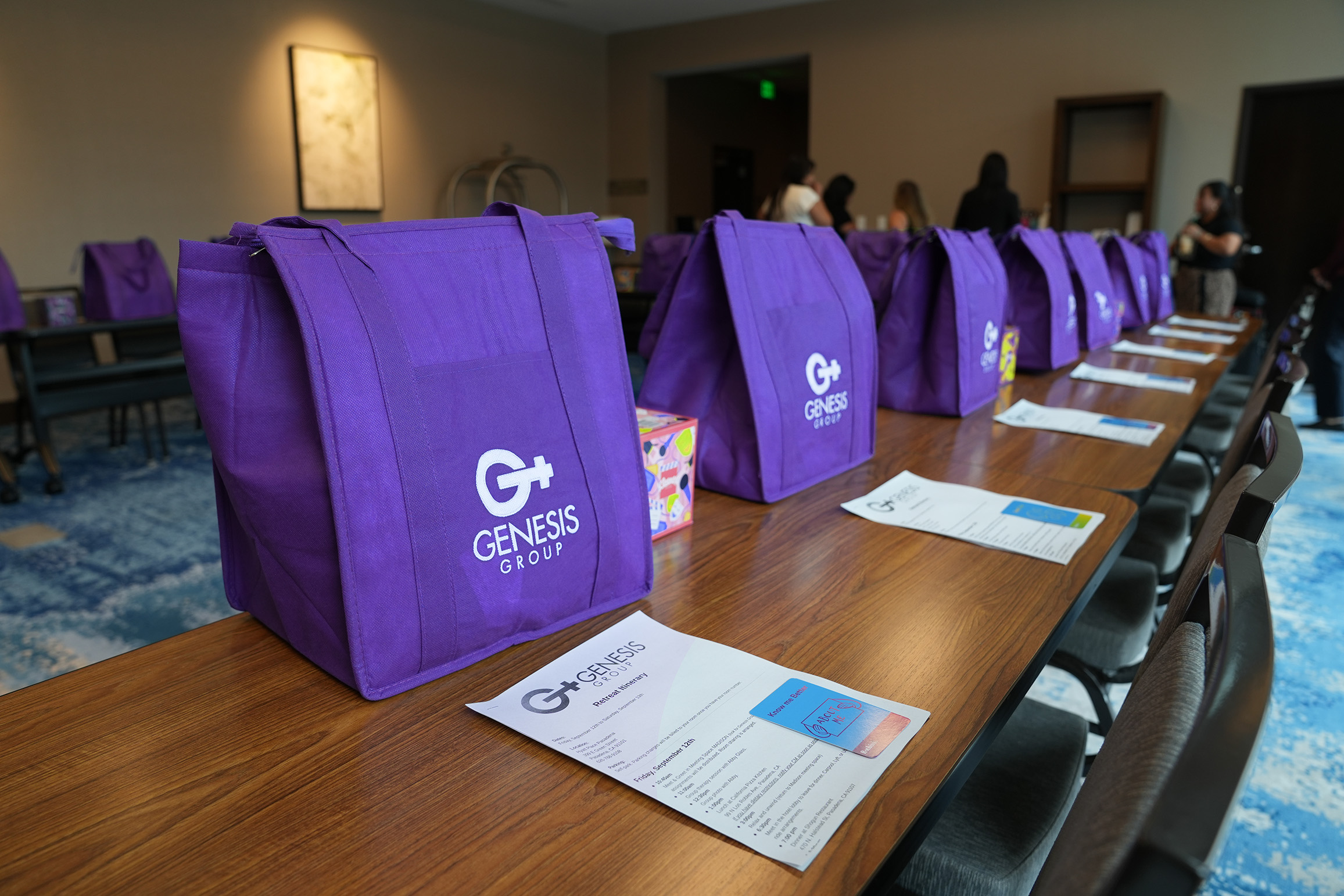 Purple bags with Genesis logo on a table, papers and people in background, indoor setting.