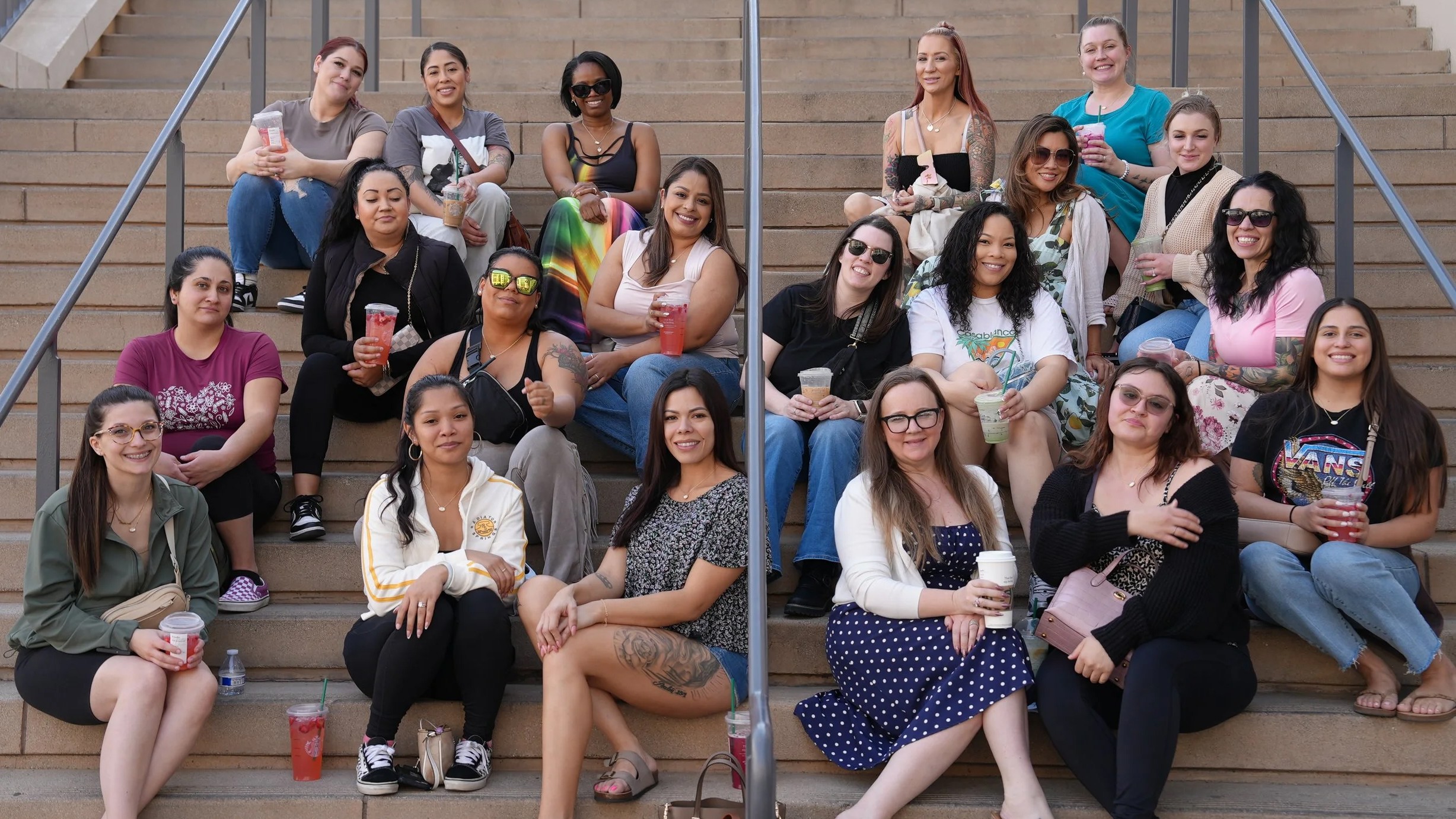 Group of women sitting on outdoor stairs, some wearing sunglasses, holding drinks, and smiling.