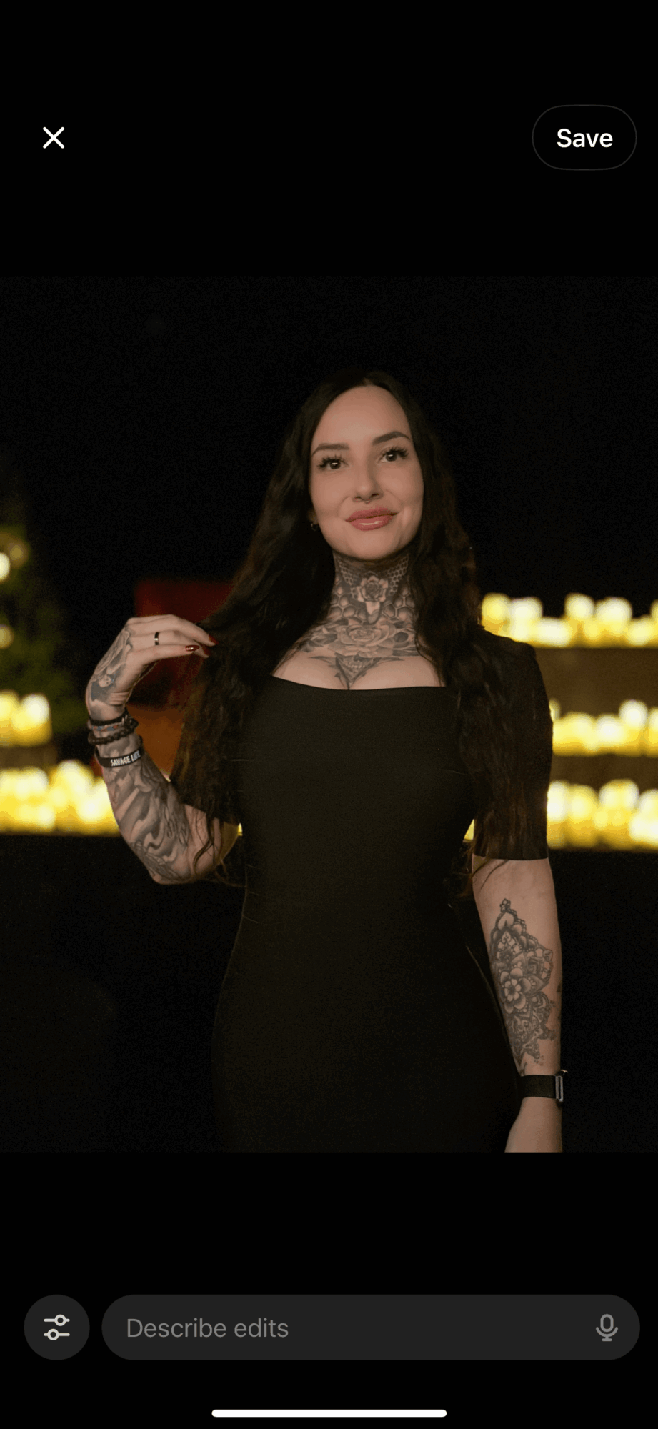 Woman in black dress with lace sleeves and tattoos, holding a drink, standing outdoors at night with lights in background.