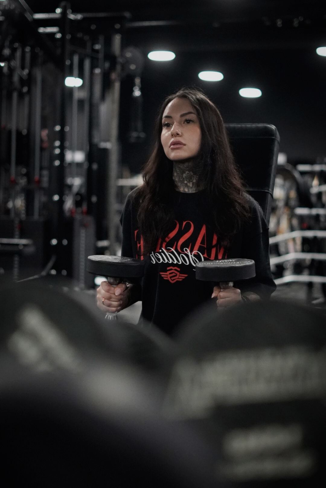 Young woman with long dark hair holding a tablet in a gym, surrounded by exercise equipment, looking serious.