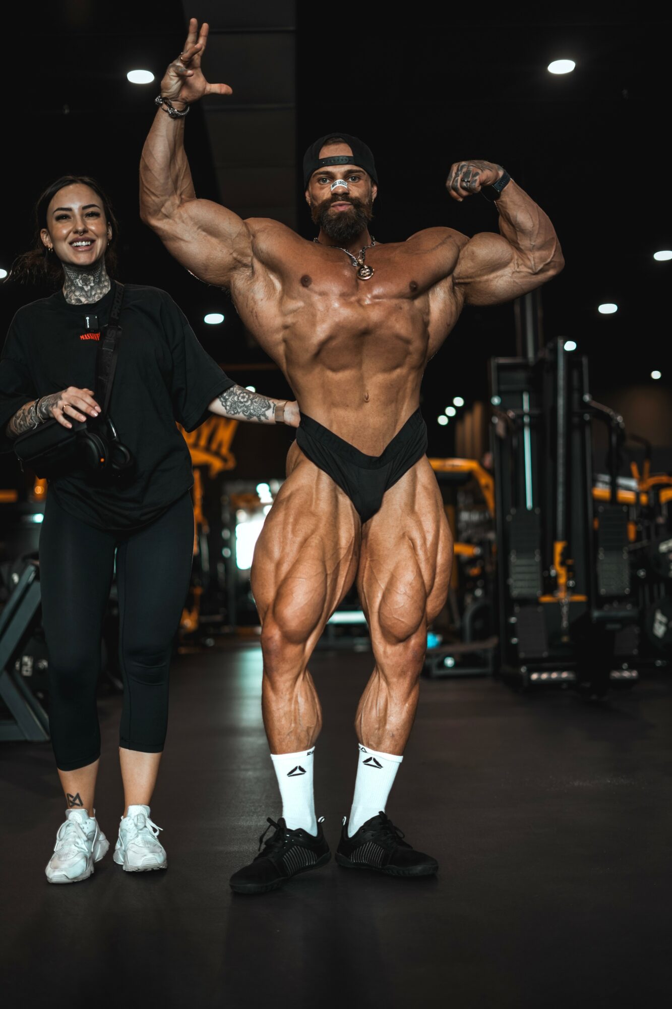 Muscular man flexing in a gym, woman standing beside him, both smiling, gym equipment in background.