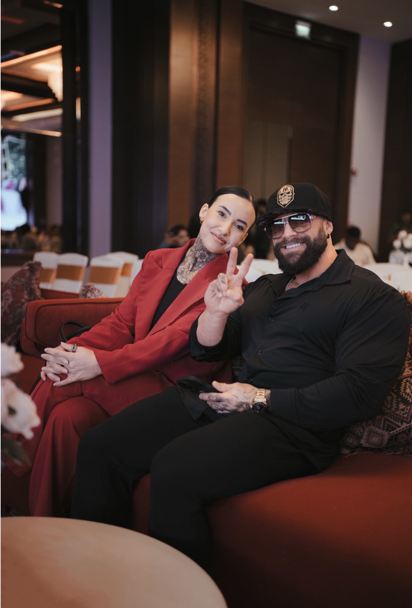 Two people sitting on a sofa, one making a peace sign, in a dimly lit indoor setting.