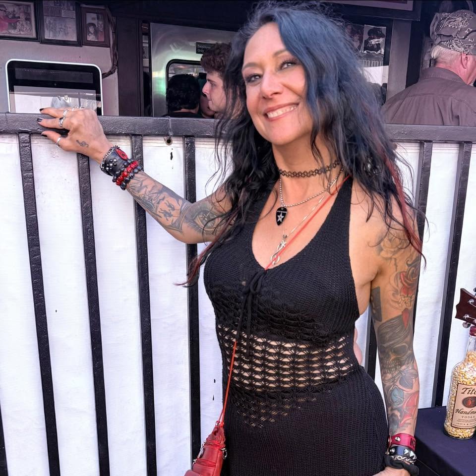Smiling woman with blue hair and tattoos, wearing a black dress, standing in front of a bar or counter.