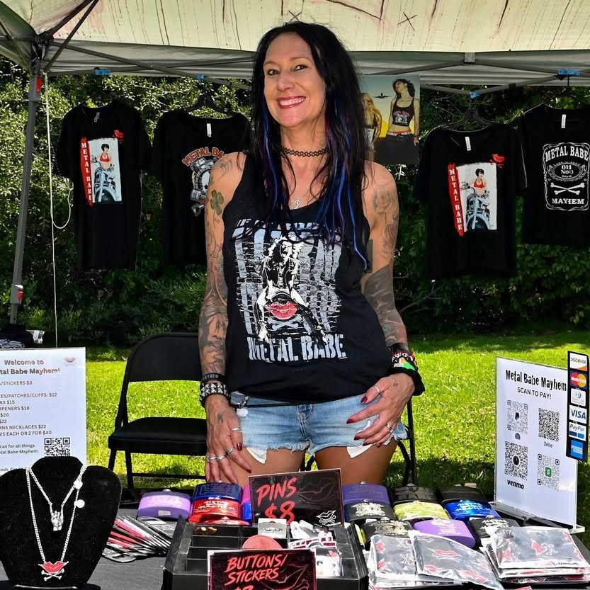Woman with long dark hair and tattoos wearing a black Metal Babe T-shirt, standing outdoors at a merchandise booth with T-shirts and accessories.