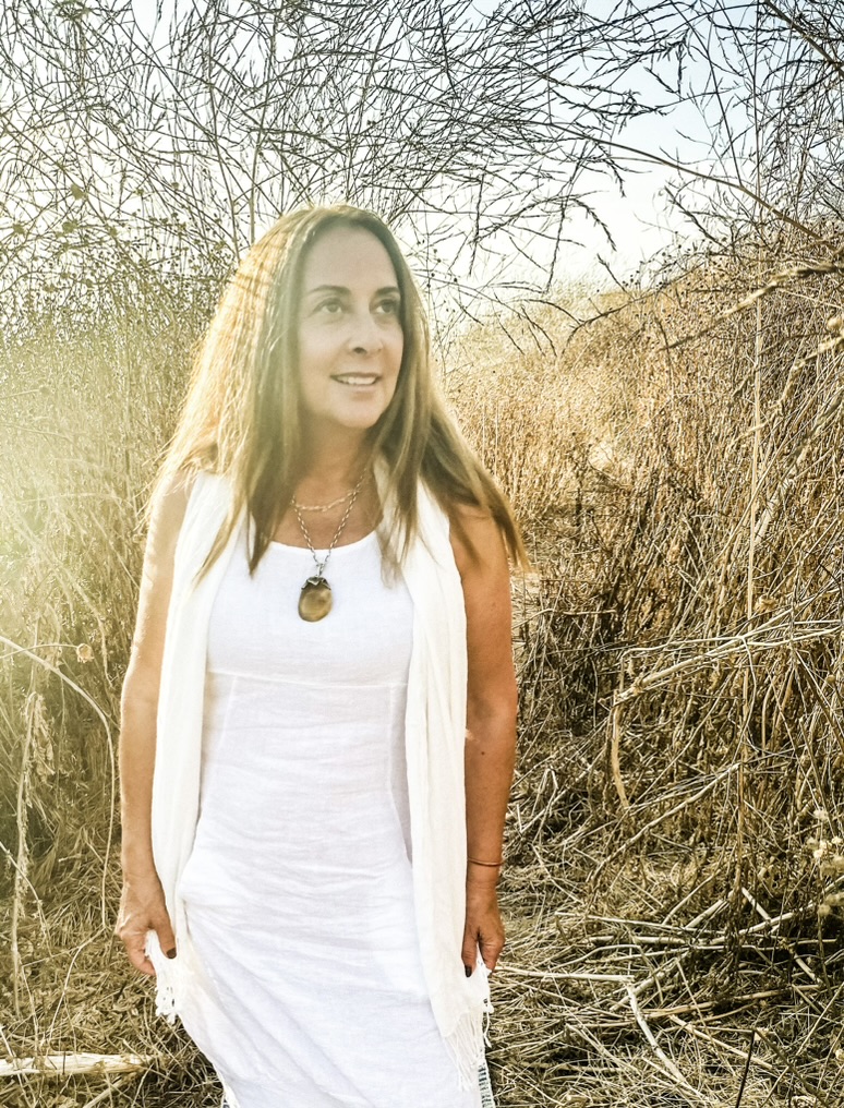 Woman with long hair in a white dress standing outdoors among dry bushes, sunlight shining behind her.