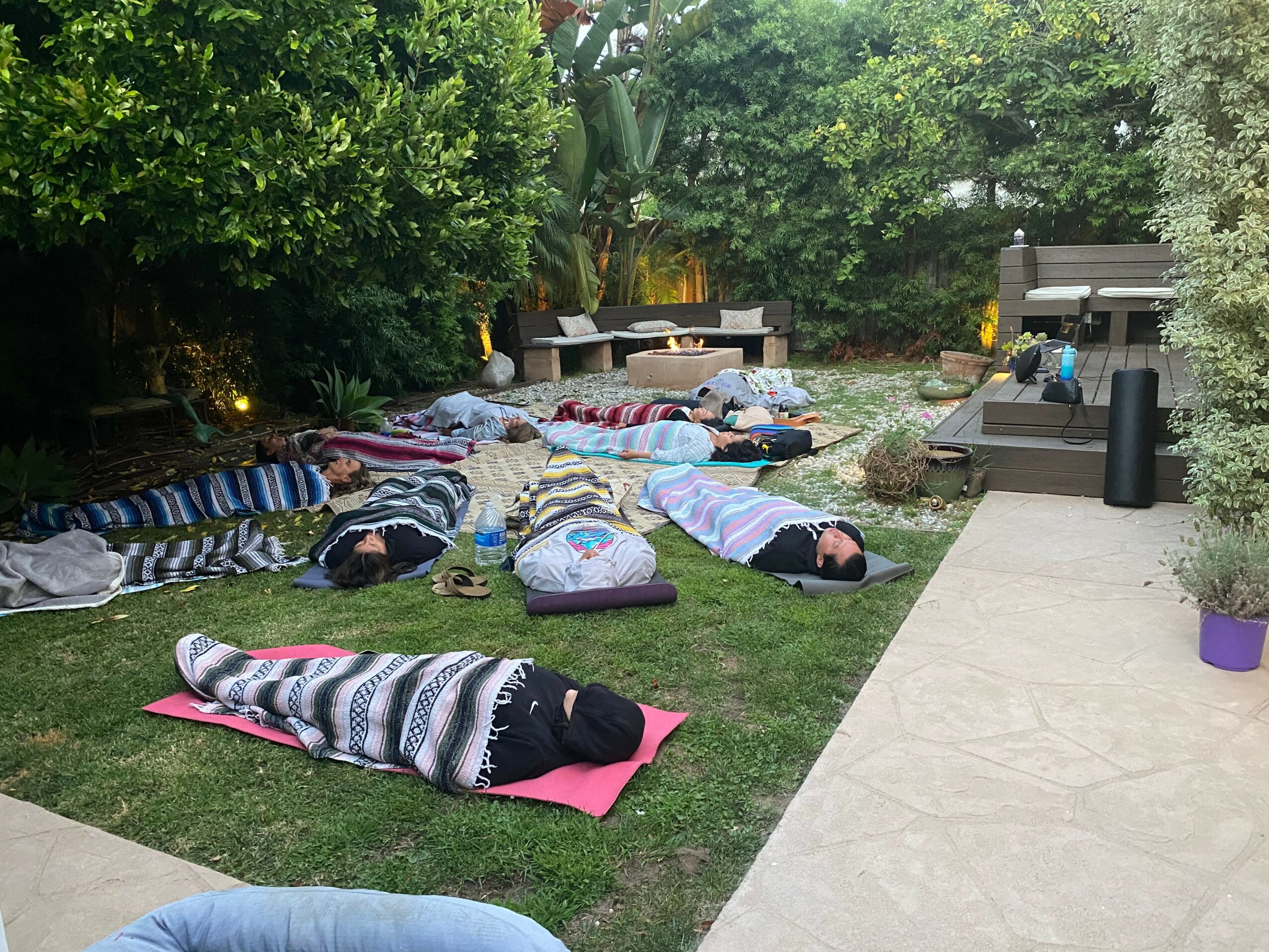 People lying on colorful blankets and mats on a grassy area outdoors, surrounded by trees and bushes, with a concrete pathway nearby.