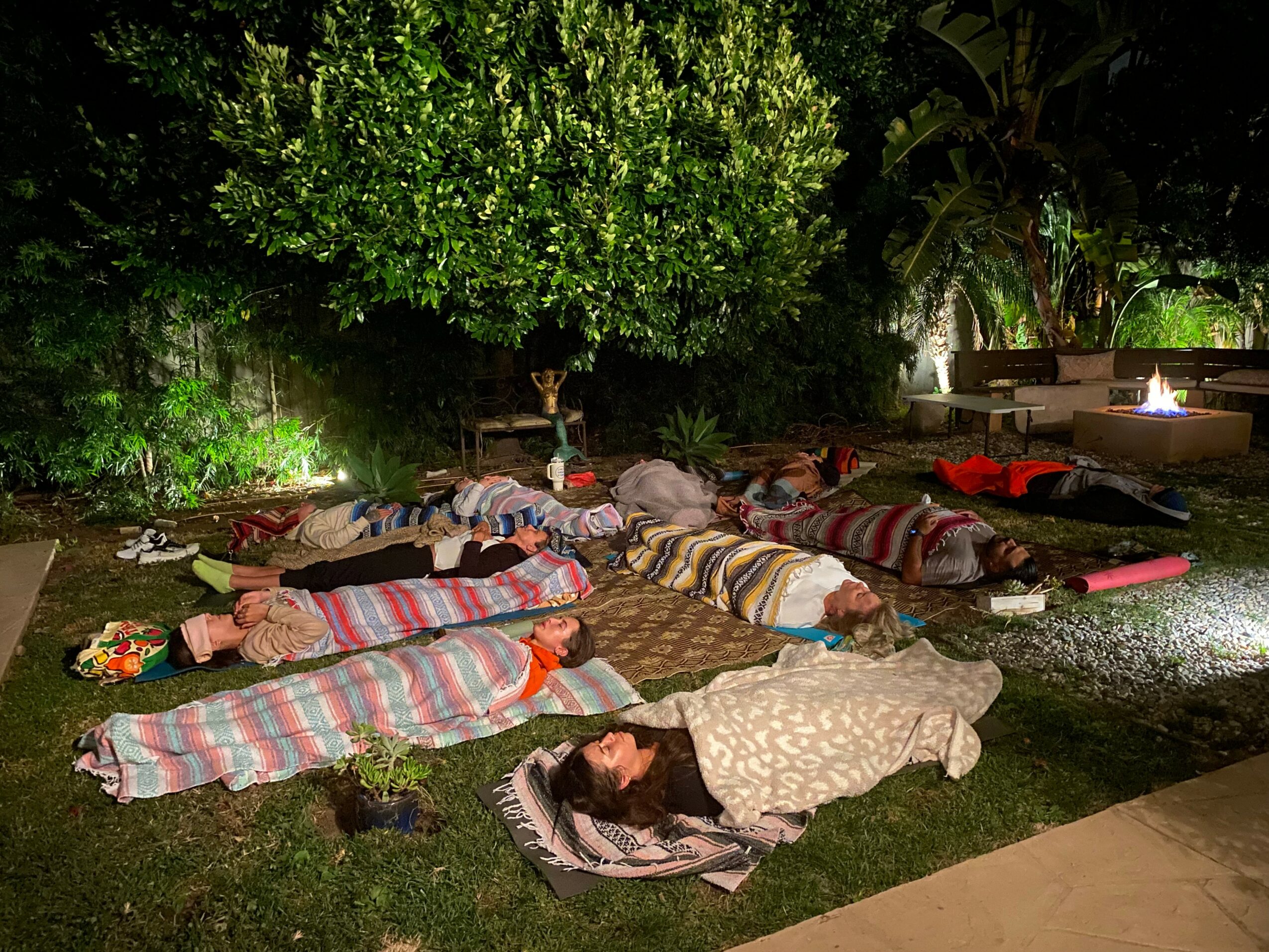 People lying on mats and blankets outdoors at night, surrounded by trees and lit candles, in a peaceful setting.