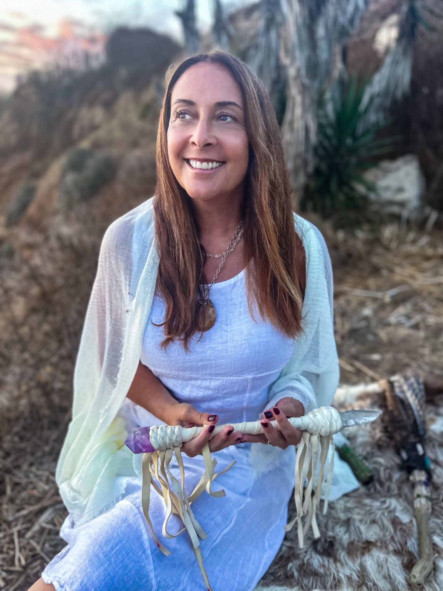 Smiling woman with long brown hair outdoors, holding a decorative staff, wearing a white dress and light shawl.