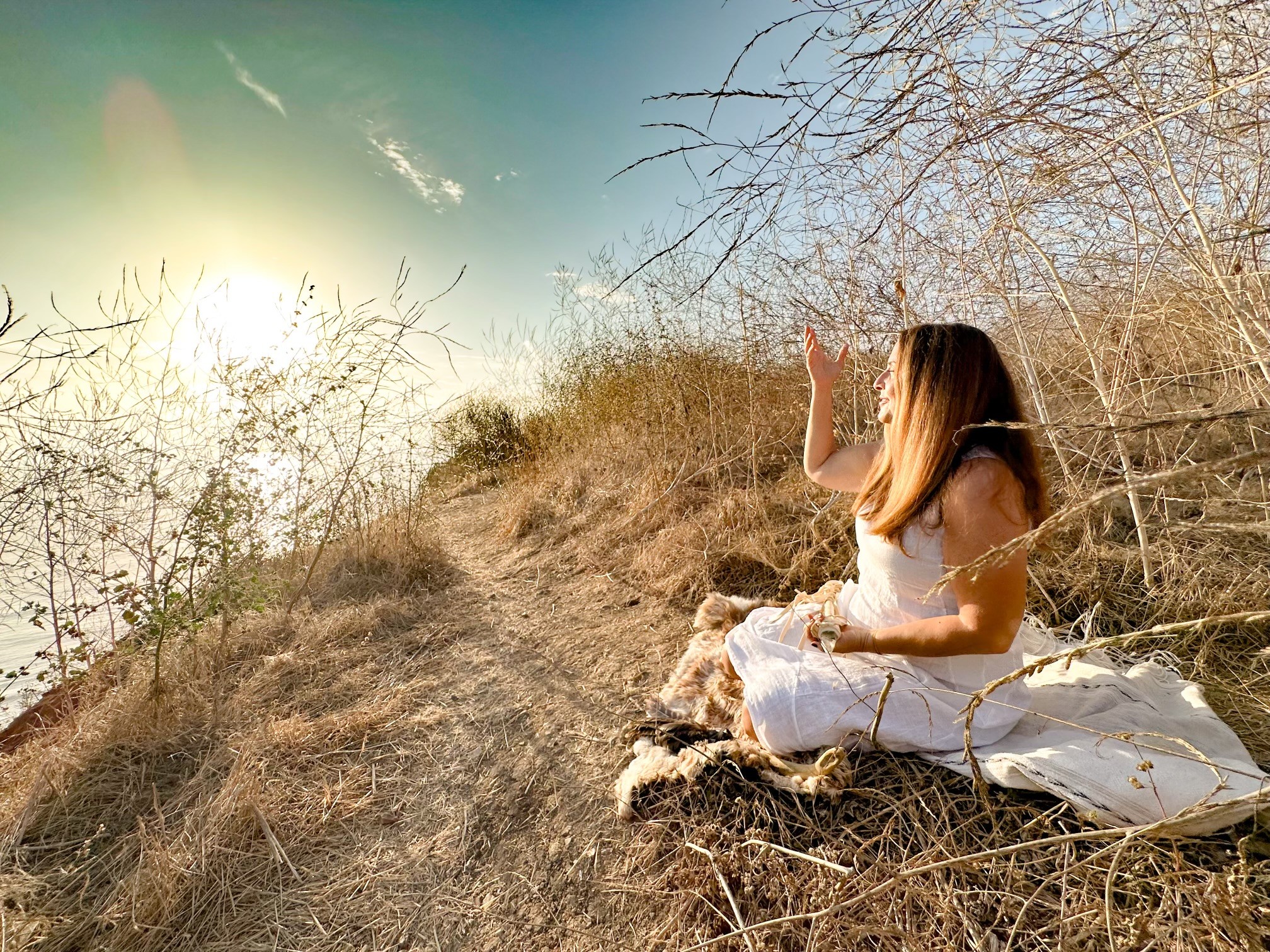 Girl in white dress sitting on dry grass hillside, waving, with leafless trees and sky in background.
