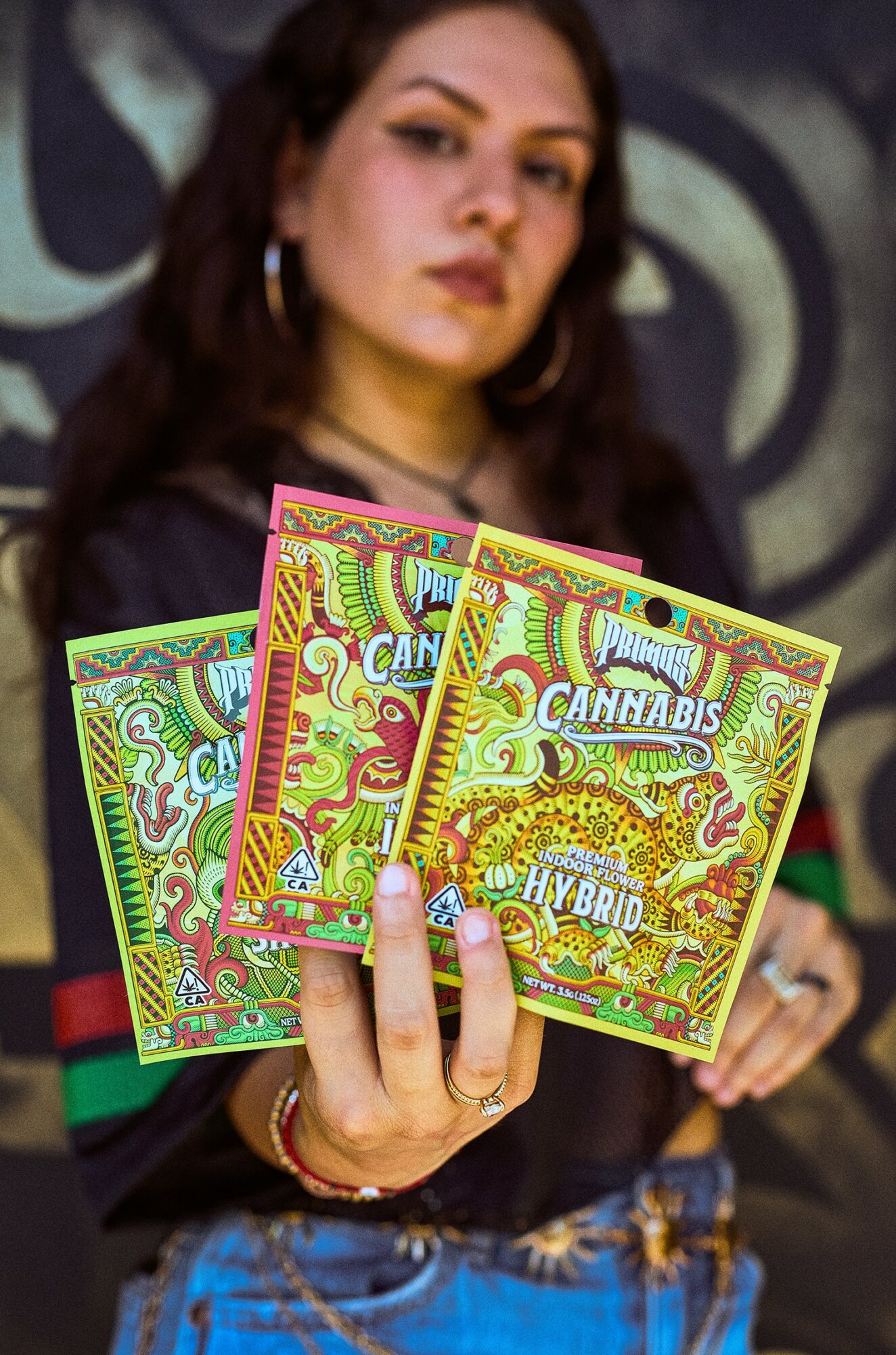 Person holding three colorful cannabis product packages, background with abstract design, wearing hoop earrings, dark hair, and a black top.