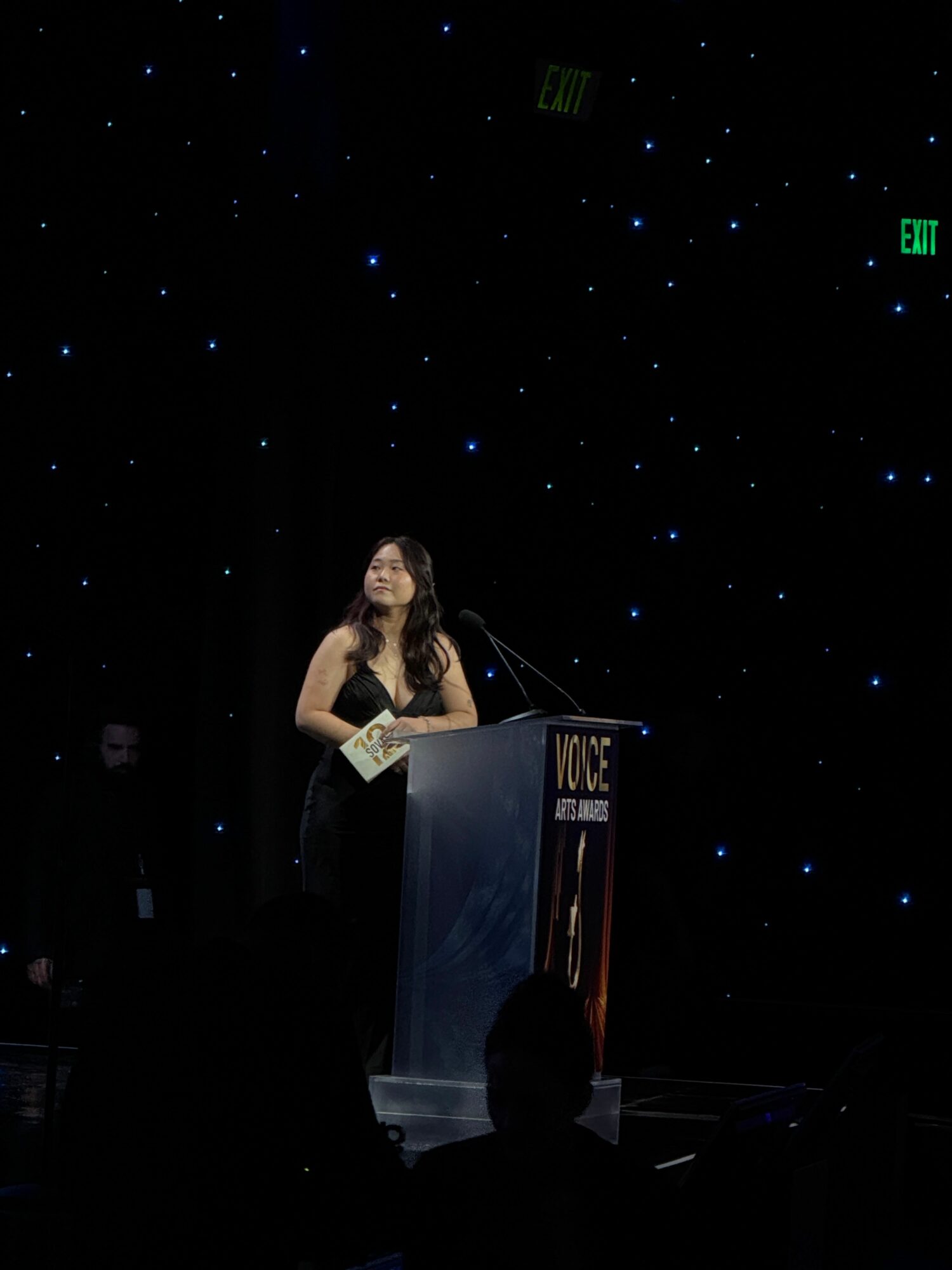 Woman speaking at a podium with a starry background, holding a microphone and notes.