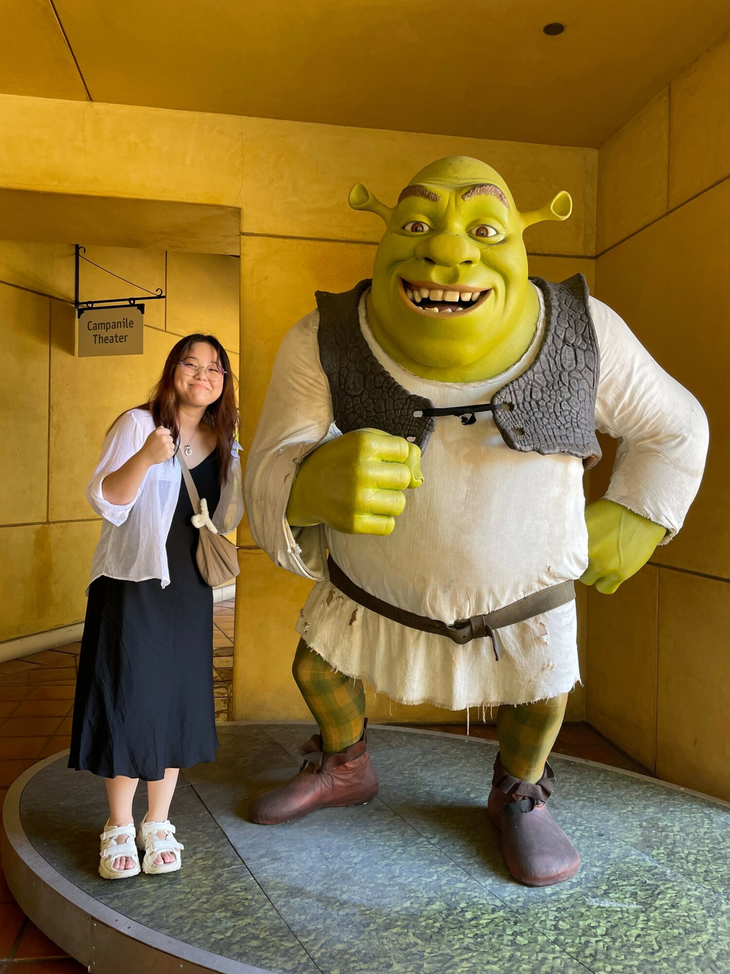 Person dressed as Shrek with green face, ears, and costume, standing with a woman in a black and white dress, indoors.