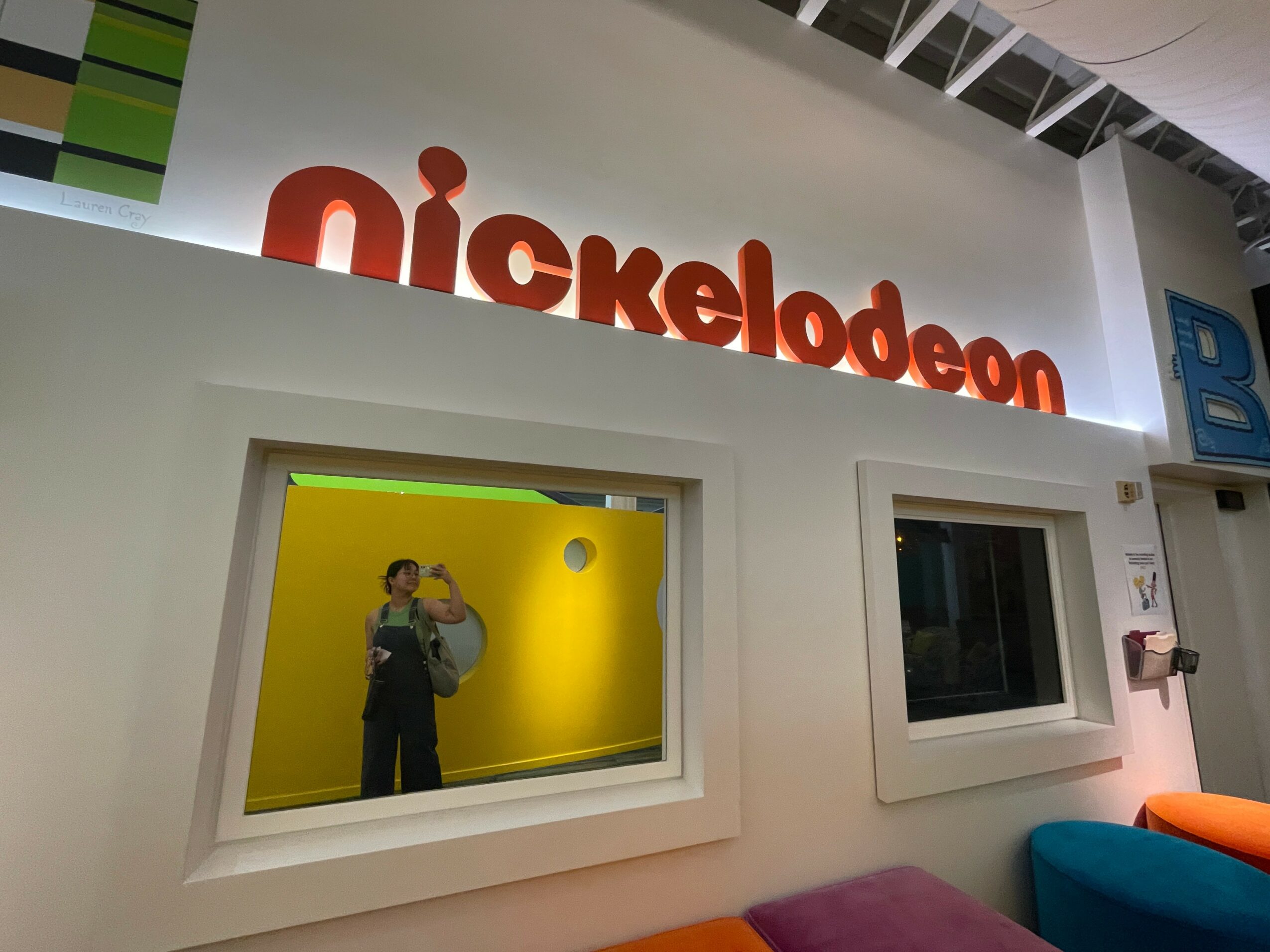 Nickelodeon sign above a window with a person taking a photo inside a yellow room, and another window with a blue and orange object nearby.