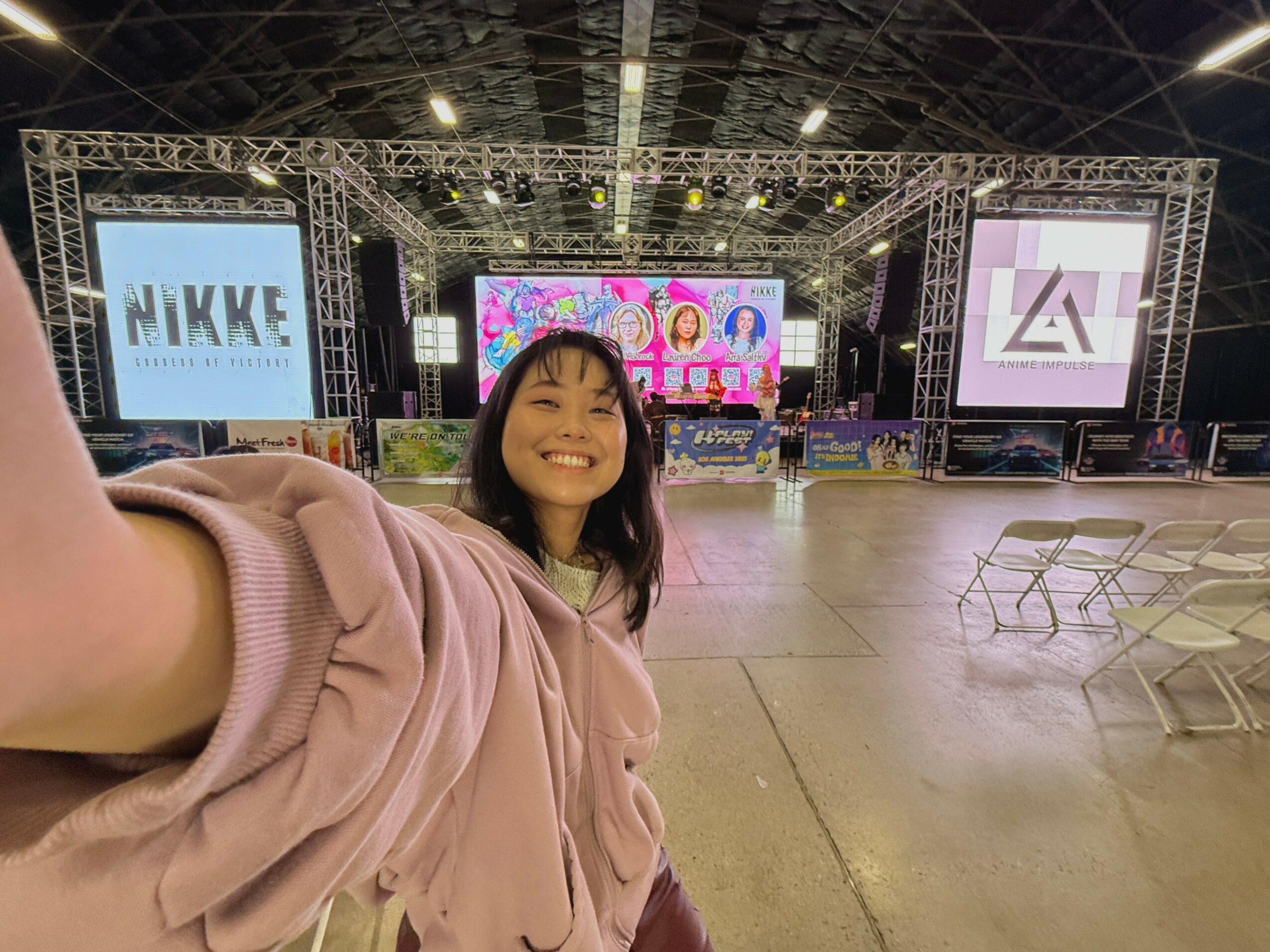 Smiling woman taking selfie in large indoor event space with stage and screens in background.