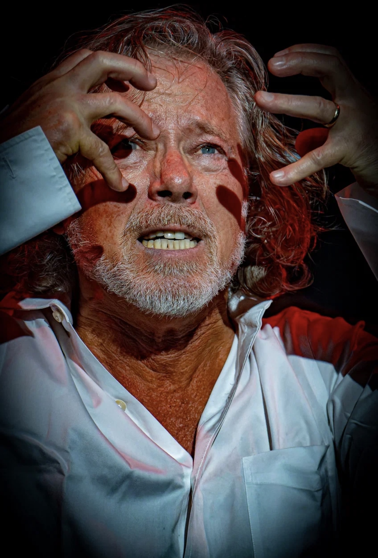 Man with gray hair and beard, wearing a white shirt, holding his head with both hands, distressed expression, dark background.