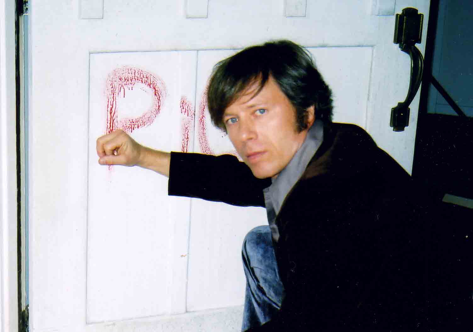Person with dark hair and black jacket drawing on a wall with red chalk, crouching near the wall.