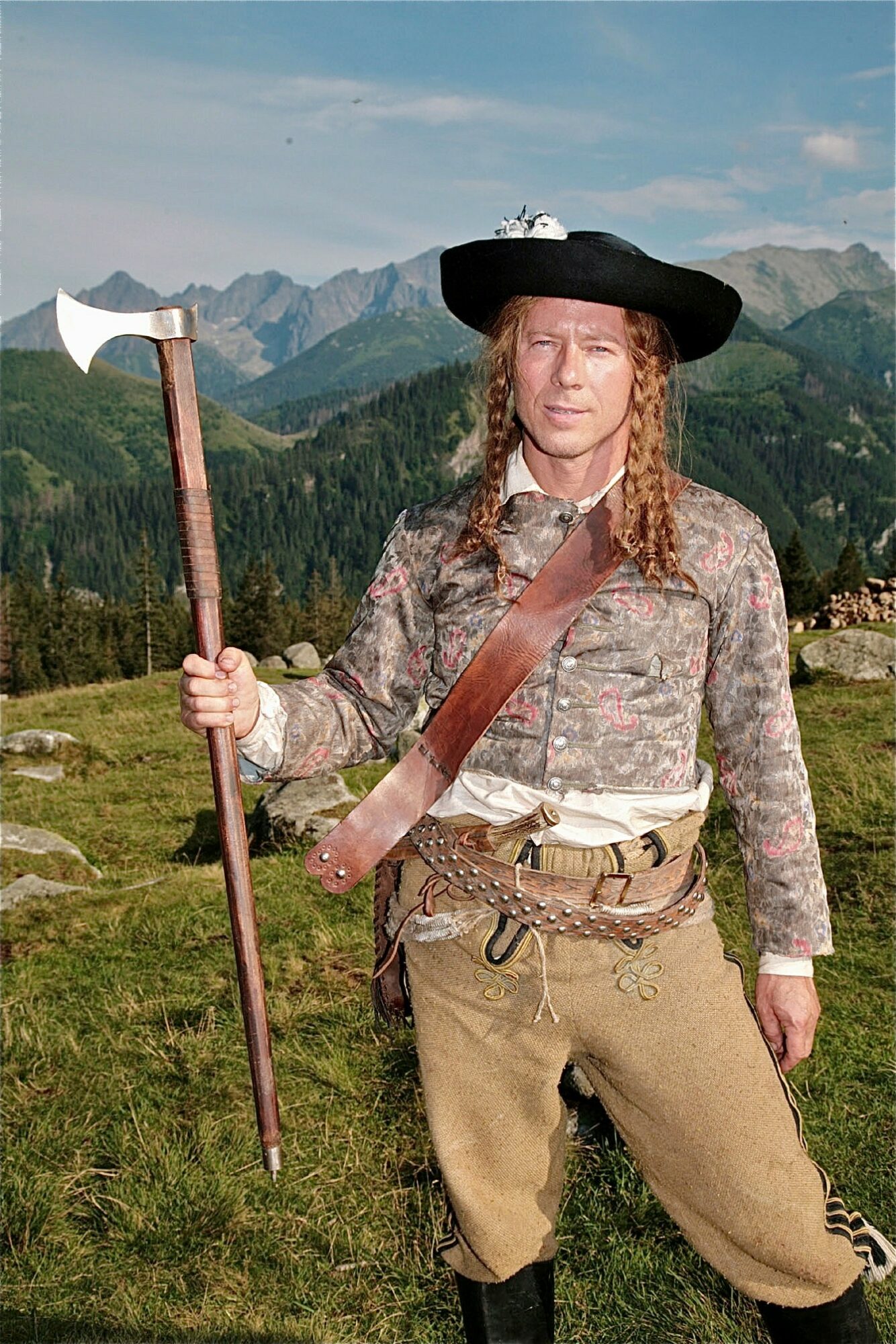 Person dressed as a historical explorer holding an axe, standing outdoors with mountains and trees in the background.