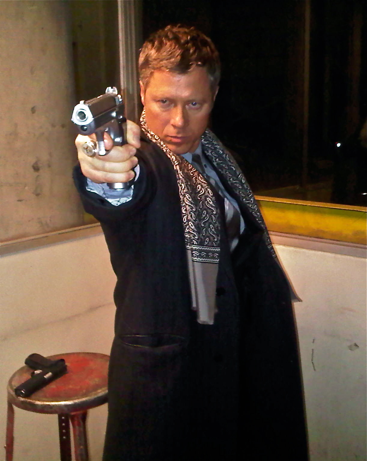 Person pointing a gun directly at the camera, indoors with a stool nearby, dark background, serious expression.