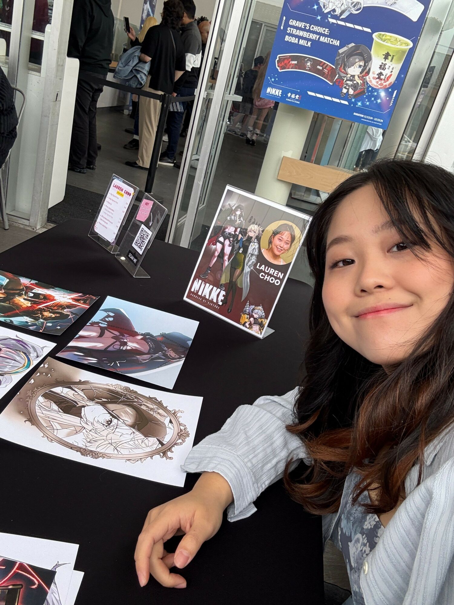 Woman with long dark hair sitting at a table with artwork and posters, in a public indoor space.