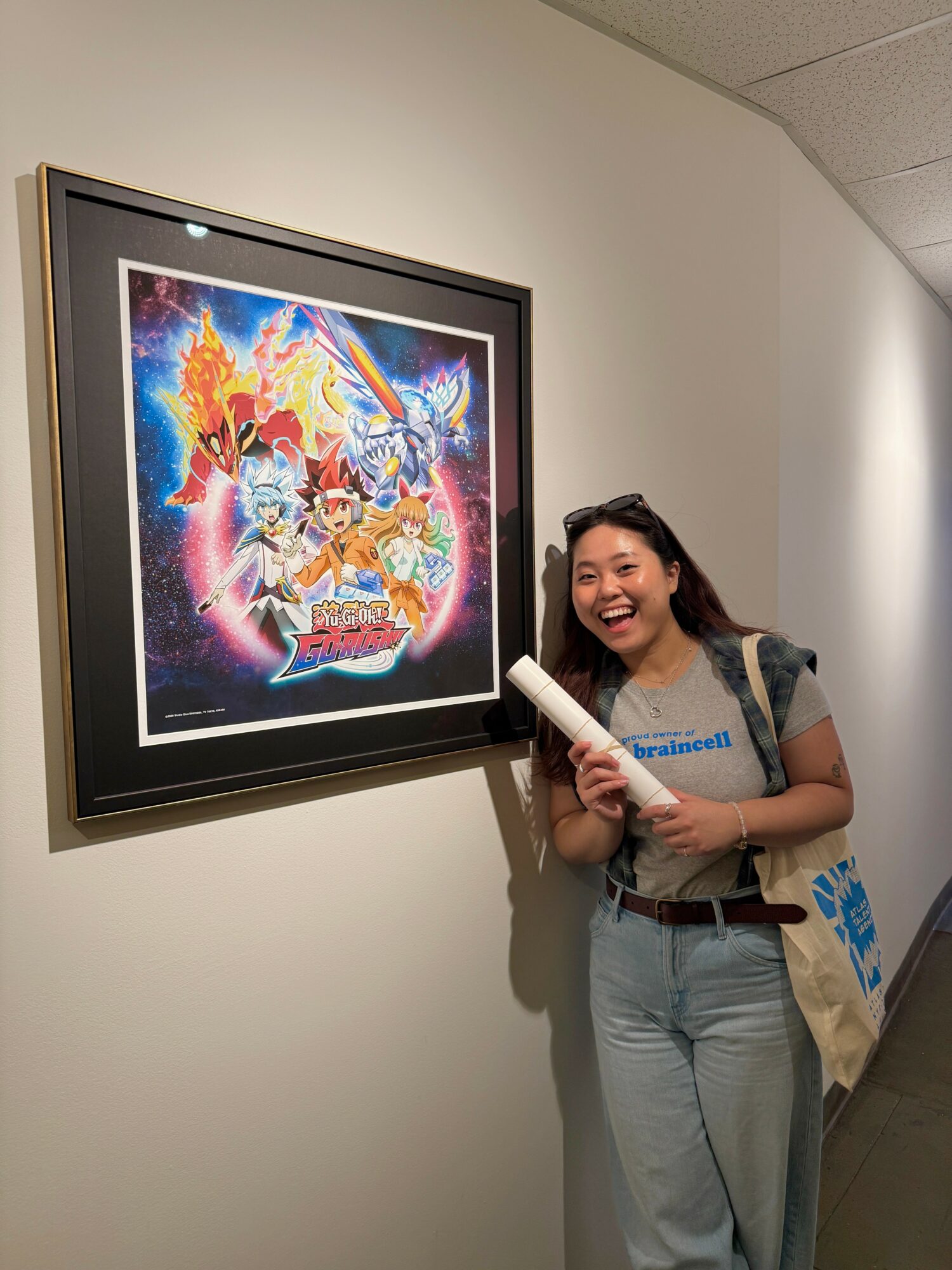 Young woman smiling and holding a rolled poster, standing next to a colorful framed artwork on a white wall.