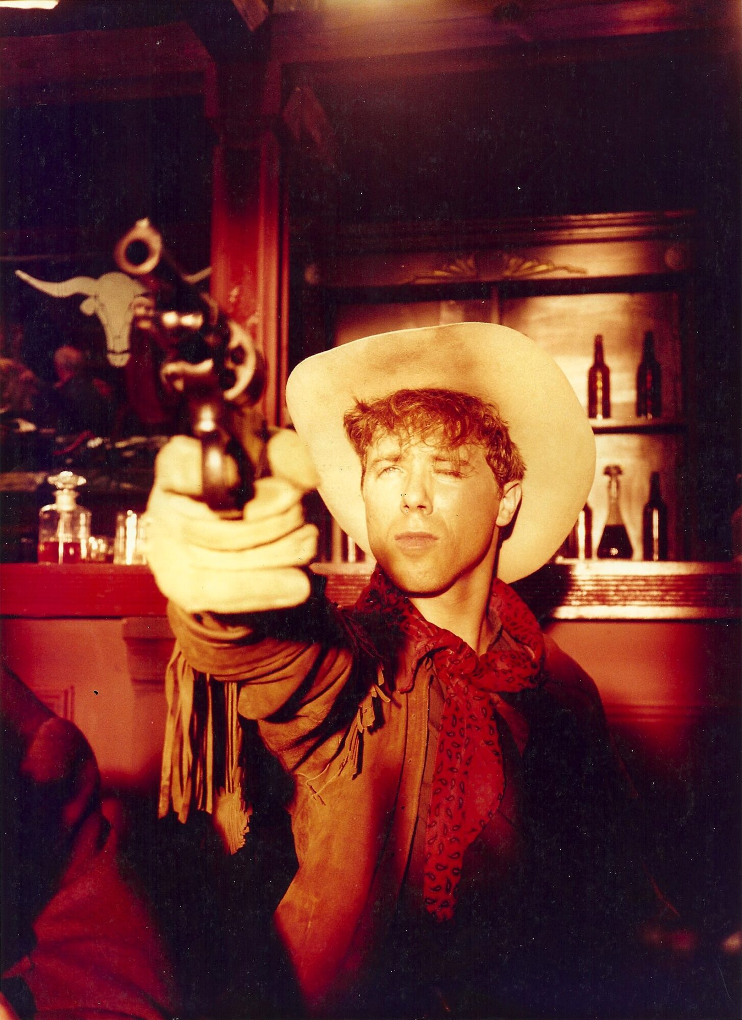 Person wearing a cowboy hat pointing a gun at the camera in a bar setting.