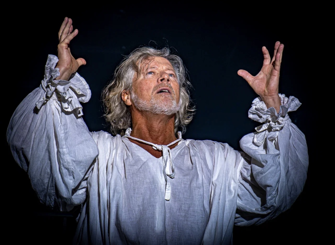 Older man with gray hair and beard, wearing a loose white shirt, raising hands with a dramatic expression against a dark background.