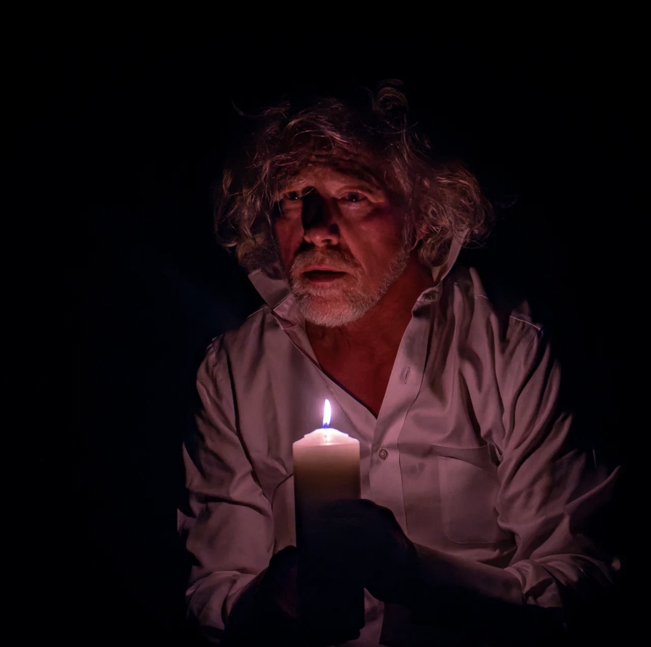 Older man with curly hair holding a lit candle in a dark environment, looking surprised or frightened.