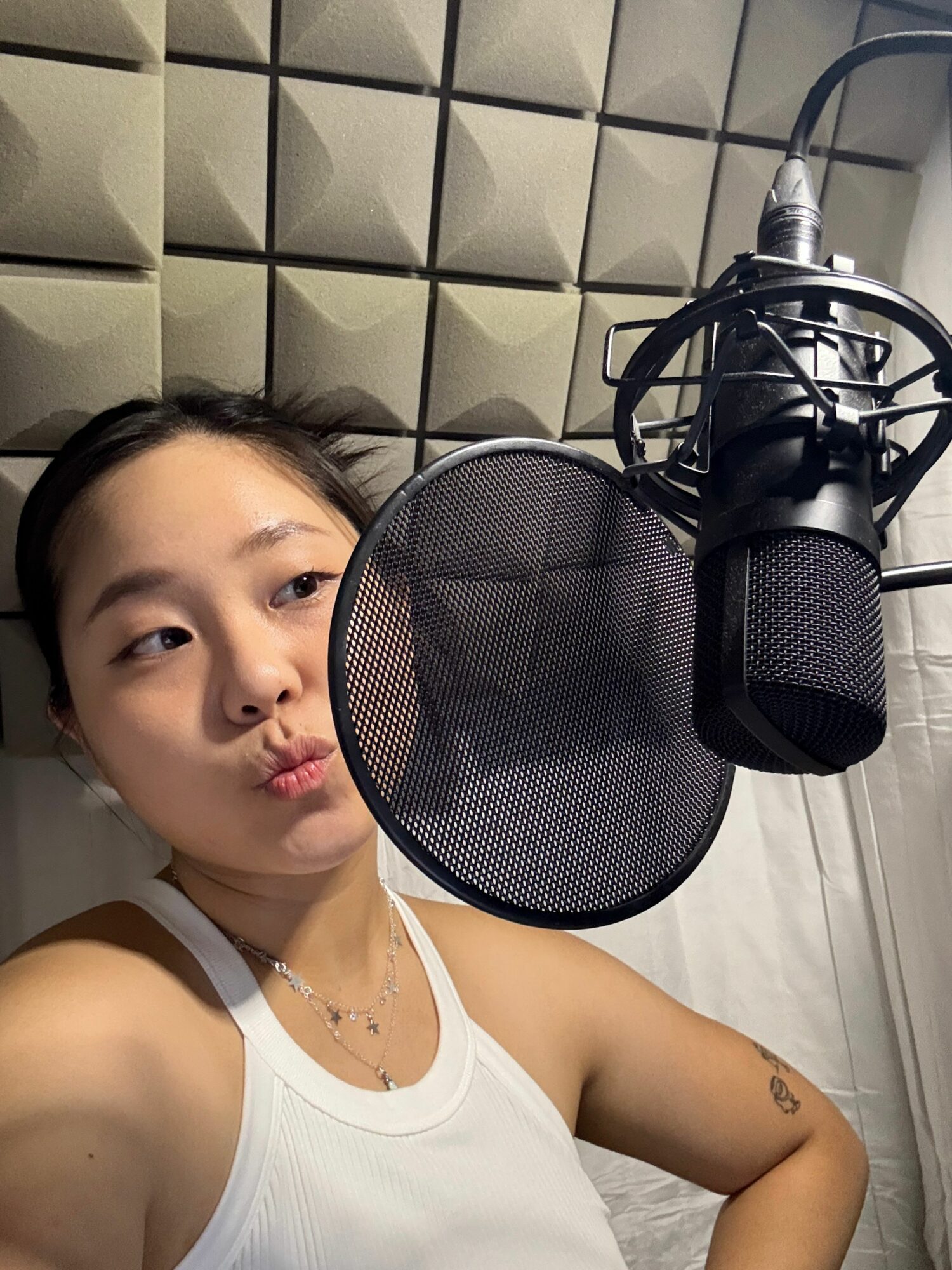 A woman lying on a bed with a microphone and pop filter overhead, wearing a white tank top, in a recording studio.