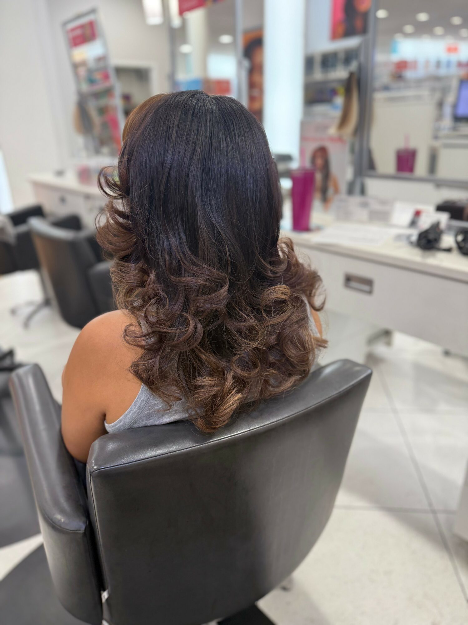 Back view of a woman with long, wavy hair sitting in a salon chair, facing a mirror, with salon equipment in background.