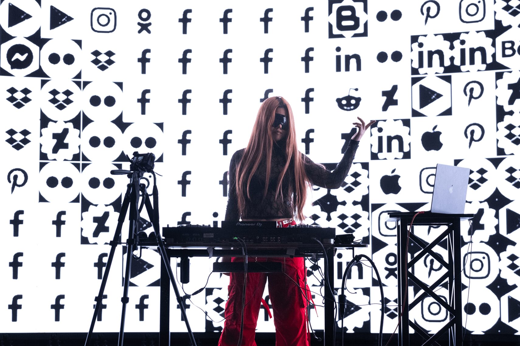 Person with long hair standing behind a table with equipment, in front of a wall with social media logos, holding a phone.