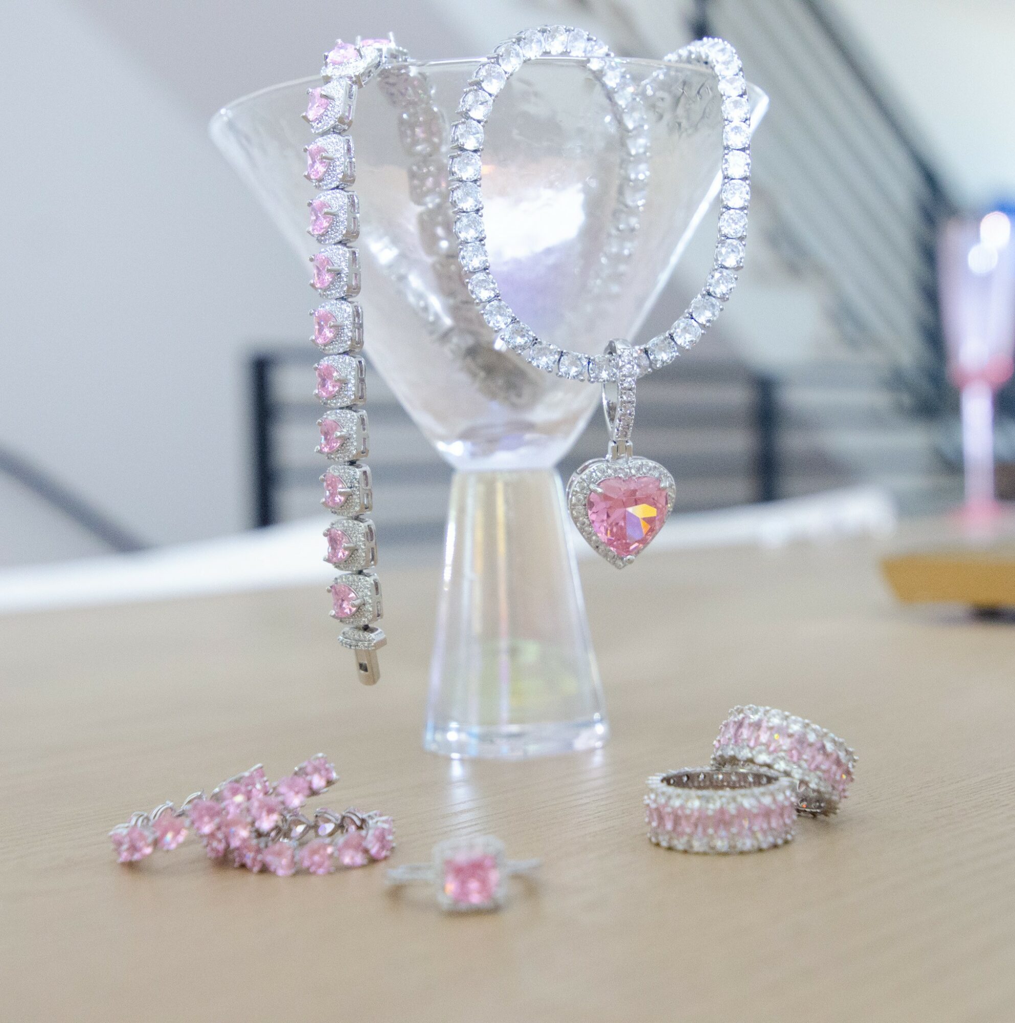 Jewelry including rings, necklaces, and a heart-shaped pendant displayed on a table with a glass and blurred background.