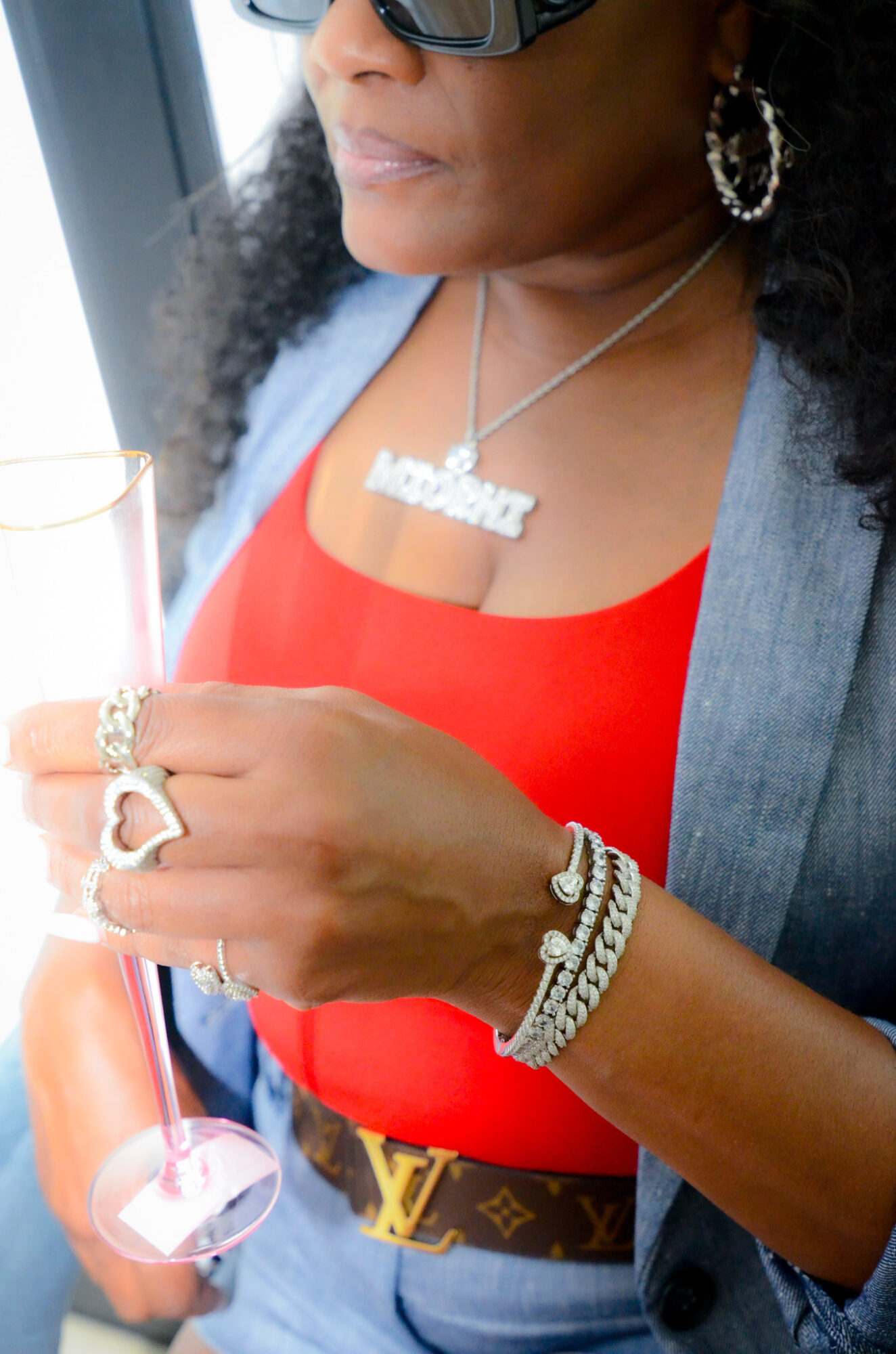 Close-up of a woman wearing sunglasses, jewelry, and a red top, holding a glass with a pink drink.