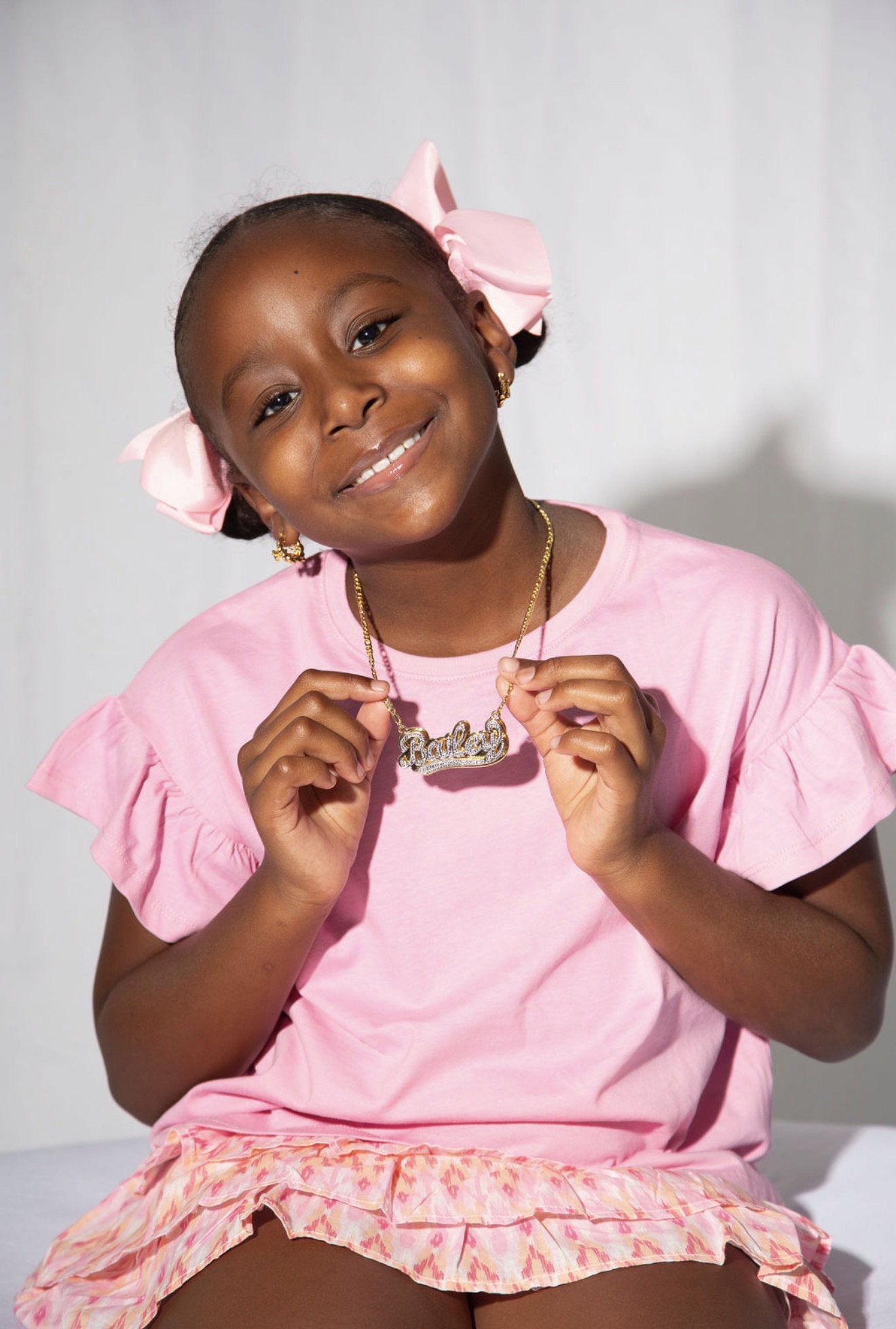 Smiling young girl with pink bows in hair, wearing pink dress and holding a necklace, against a plain background.