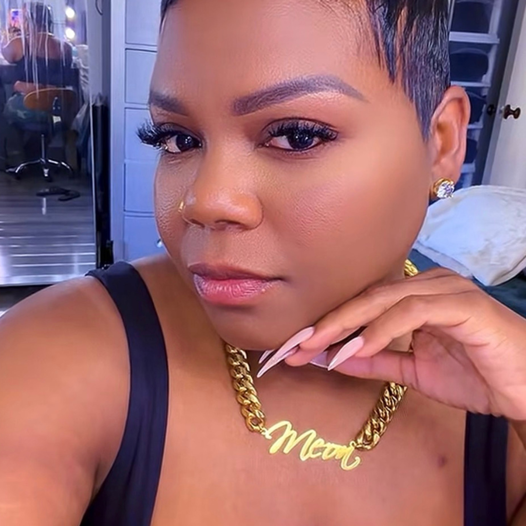 Close-up of a woman with short hair, wearing a gold necklace with the word 'Mew' and a black top, indoors.