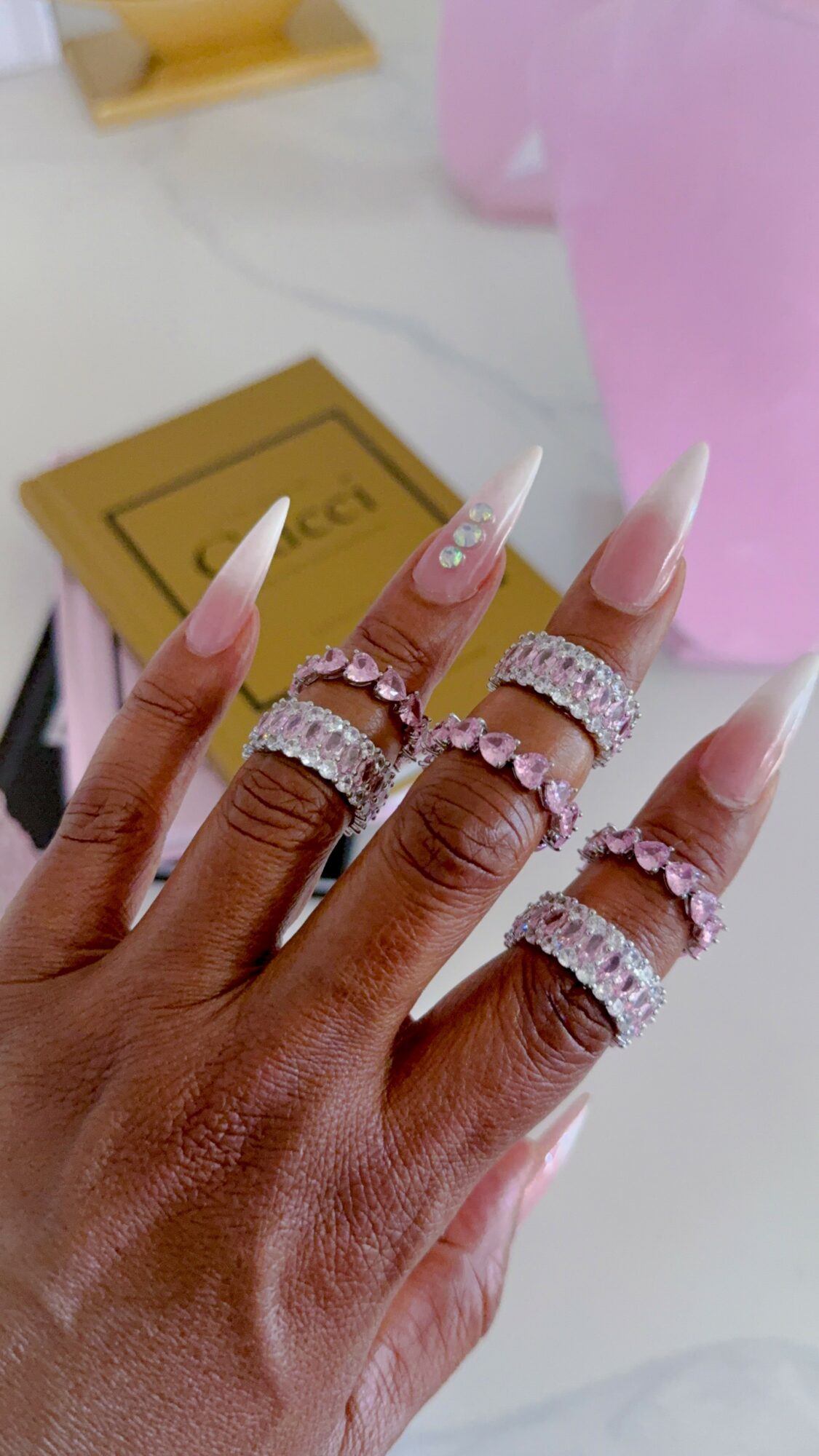 Hand with long, pointed, pink and white nails, wearing multiple rings, resting on a surface with a gold box nearby.