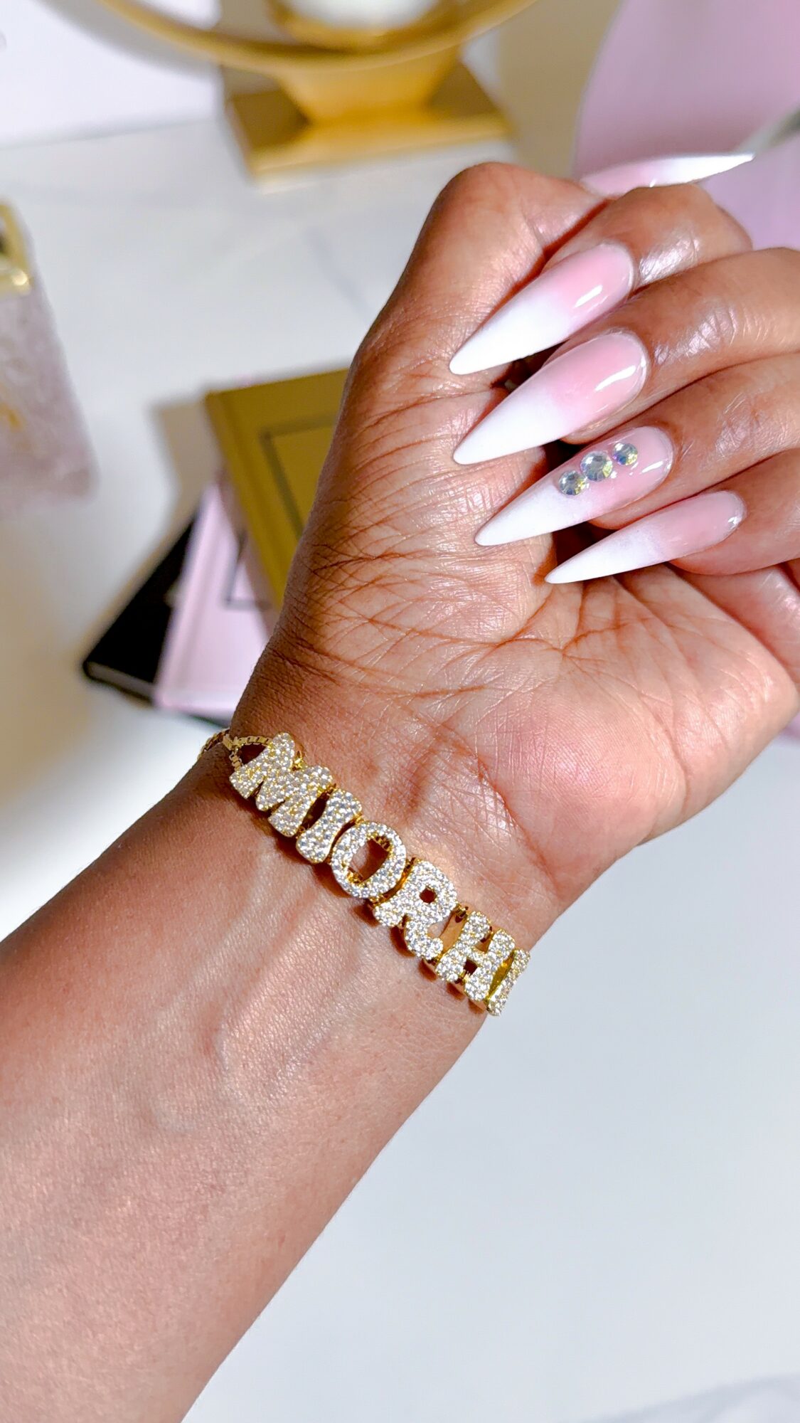 Hand with long, white, decorated nails and a gold bracelet spelling 'MUM' on wrist.
