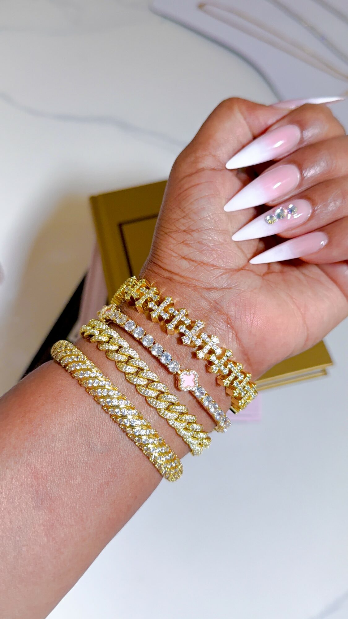 Hand with five gold and pink bracelets, long white nails with rhinestones, against a blurred background.