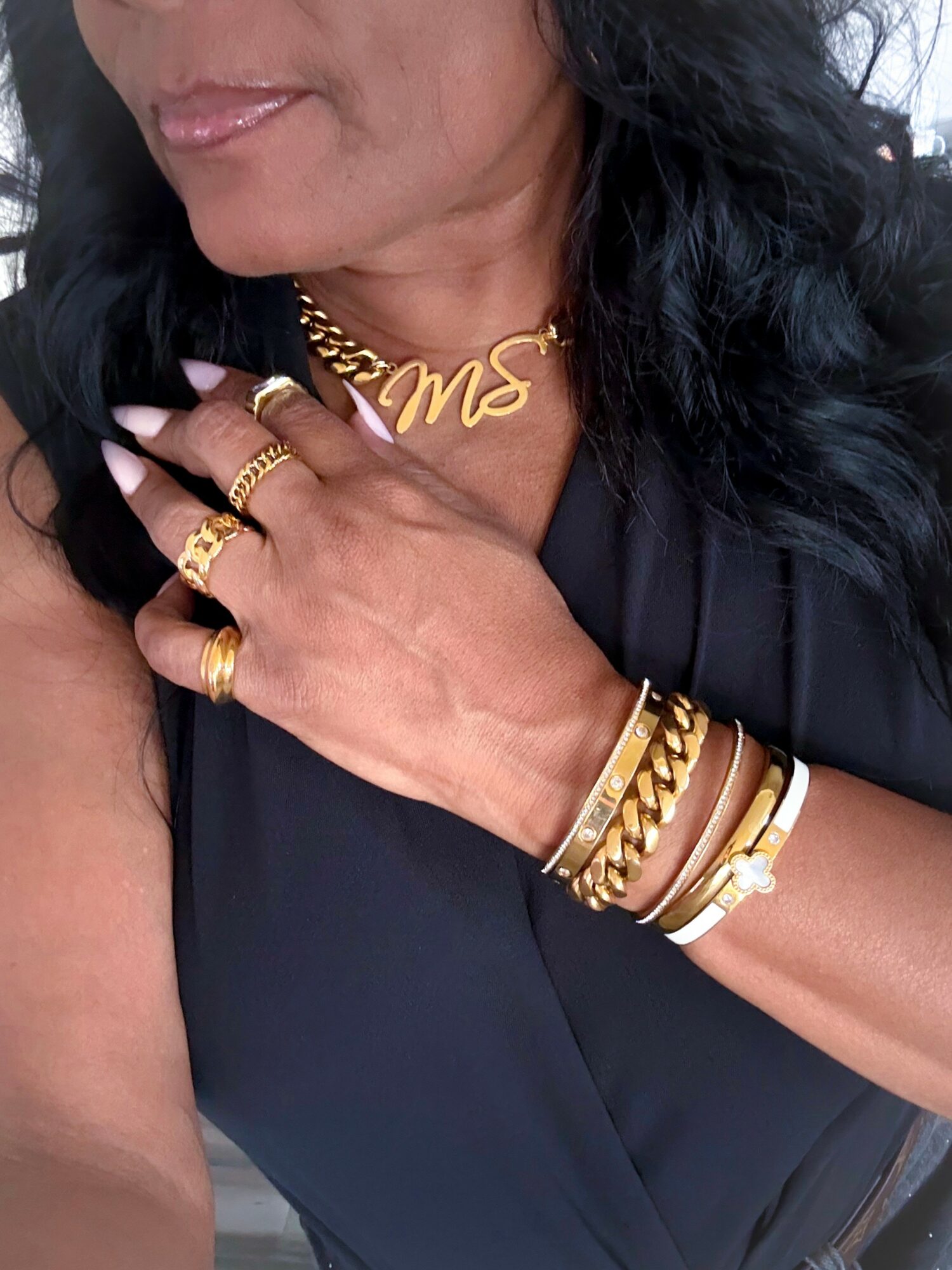 Close-up of a woman wearing gold jewelry, including rings, bracelets, and a necklace, with her hand on her chest.