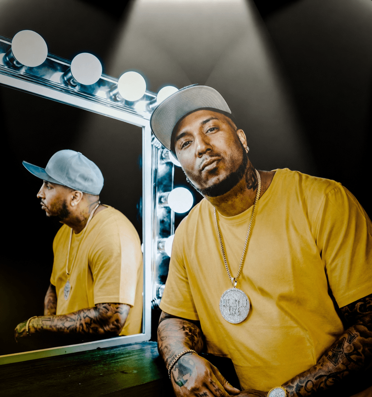 Man with tattoos wearing a yellow shirt and gray cap, standing in front of a mirror with lights, reflection visible.